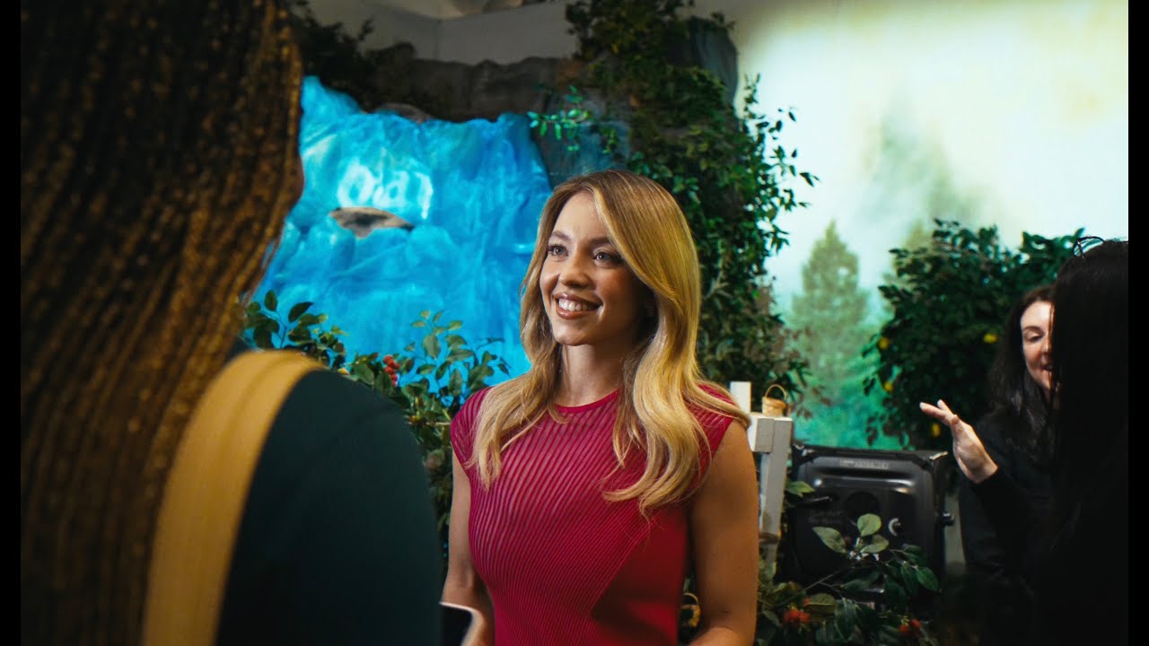 Sydney Sweeney in a red dress smiles at guests in front of a painted forest and waterfall set