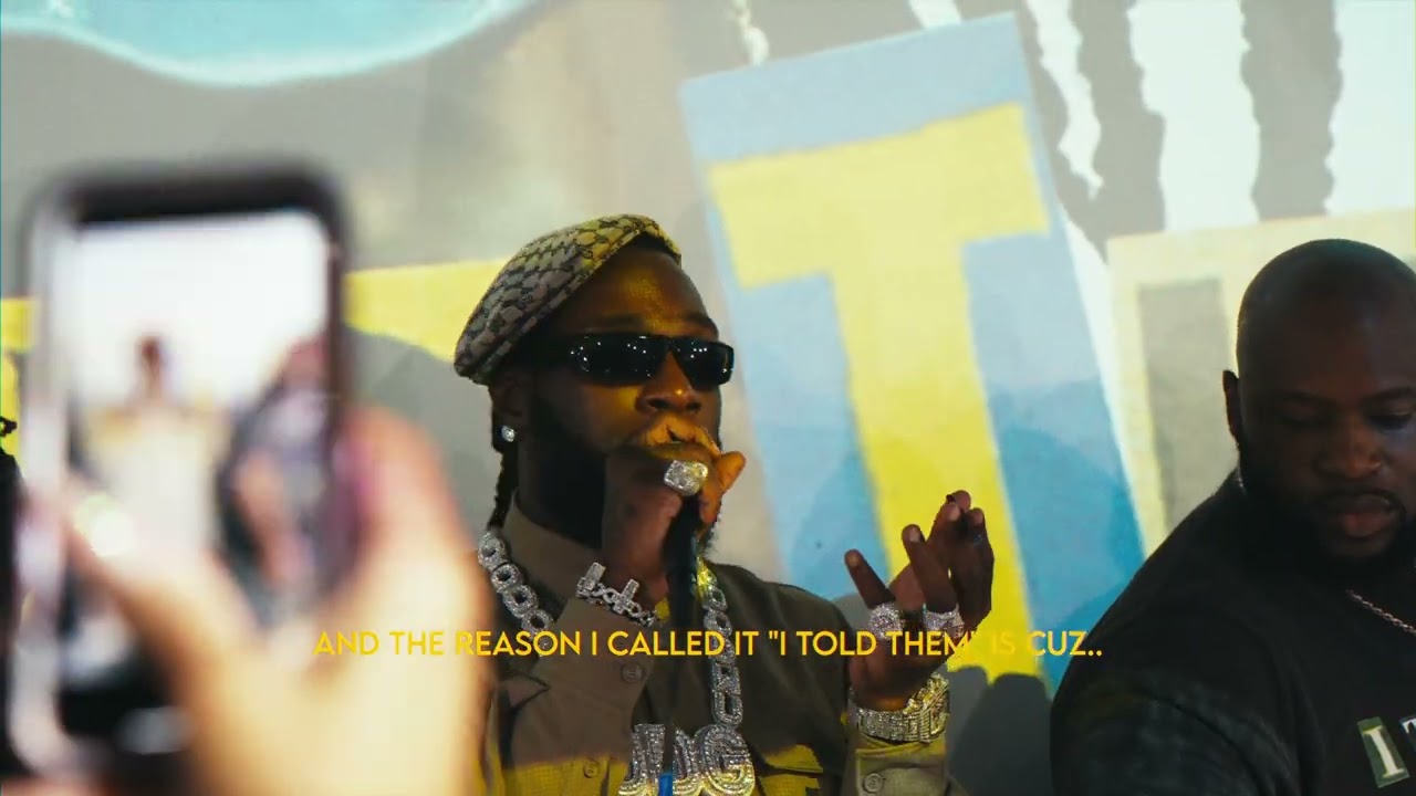Burna Boy in sunglasses and a patterned cap speaks into a microphone as phones film him