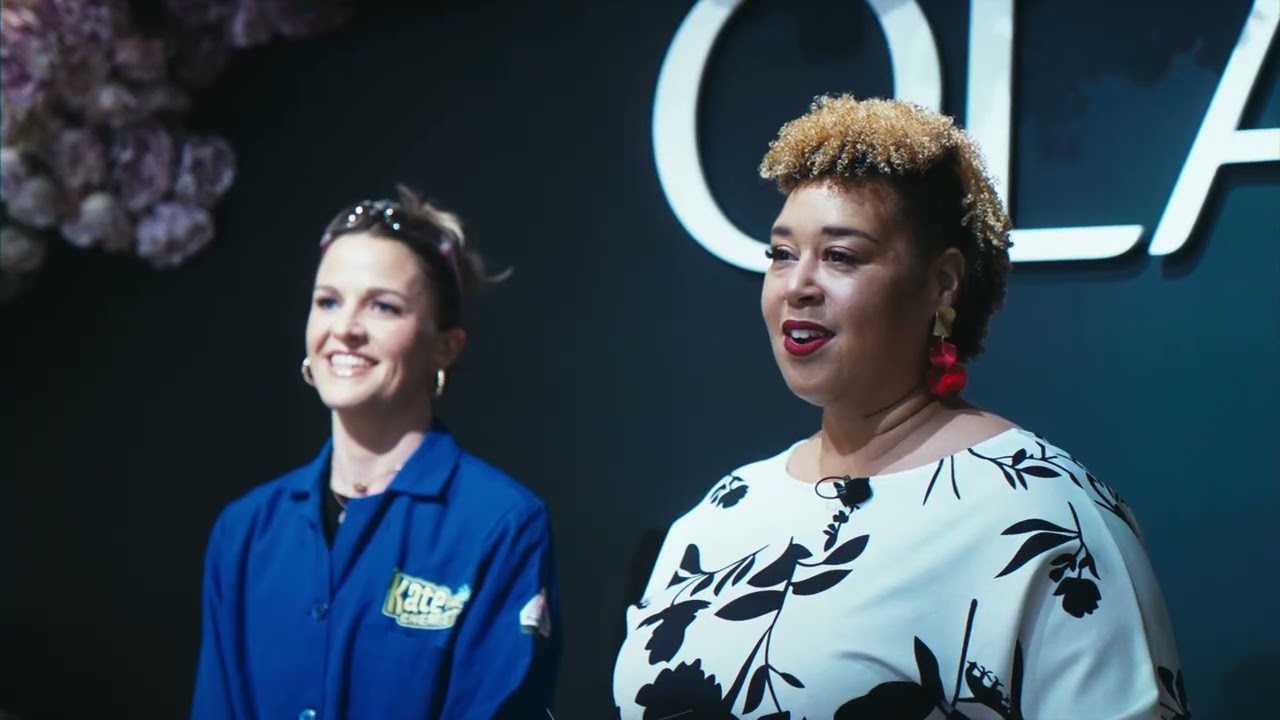 Two smiling presenters in front of a dark wall with the OLAY logo and hanging flowers