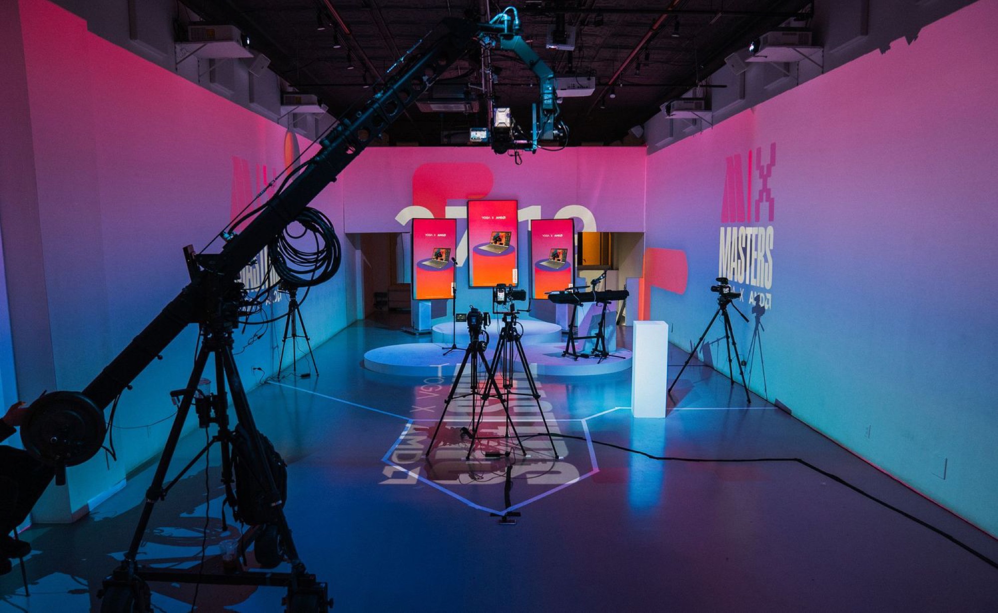 Camera jib and tripods aimed at three lit product display screens on the Lenovo x AMD Masters stage, set under pink and blue gradient lighting