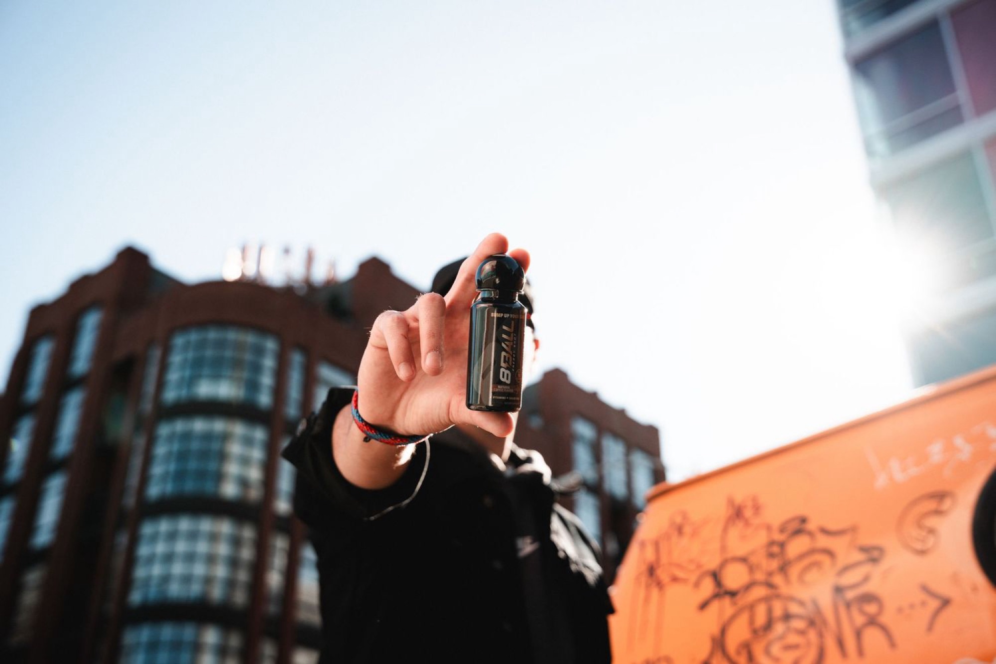 Hand holding an 8Ball Energy shot up to the sky between city buildings and an orange graffiti wall