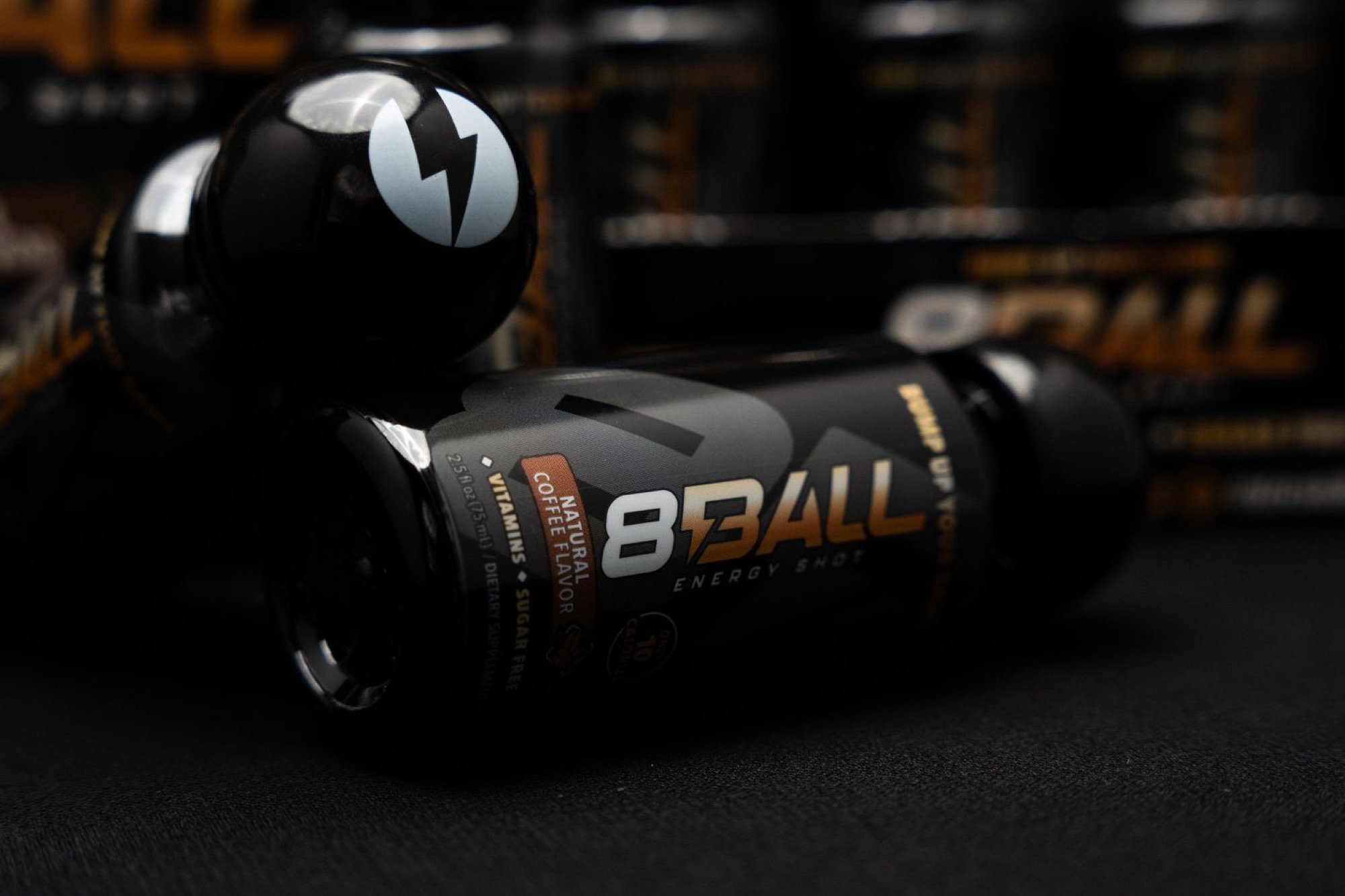Two 8Ball Energy Shot bottles on a dark surface, the front one on its side with its black and orange label for the coffee flavor lit, rows of branded 8Ball cartons blurred behind