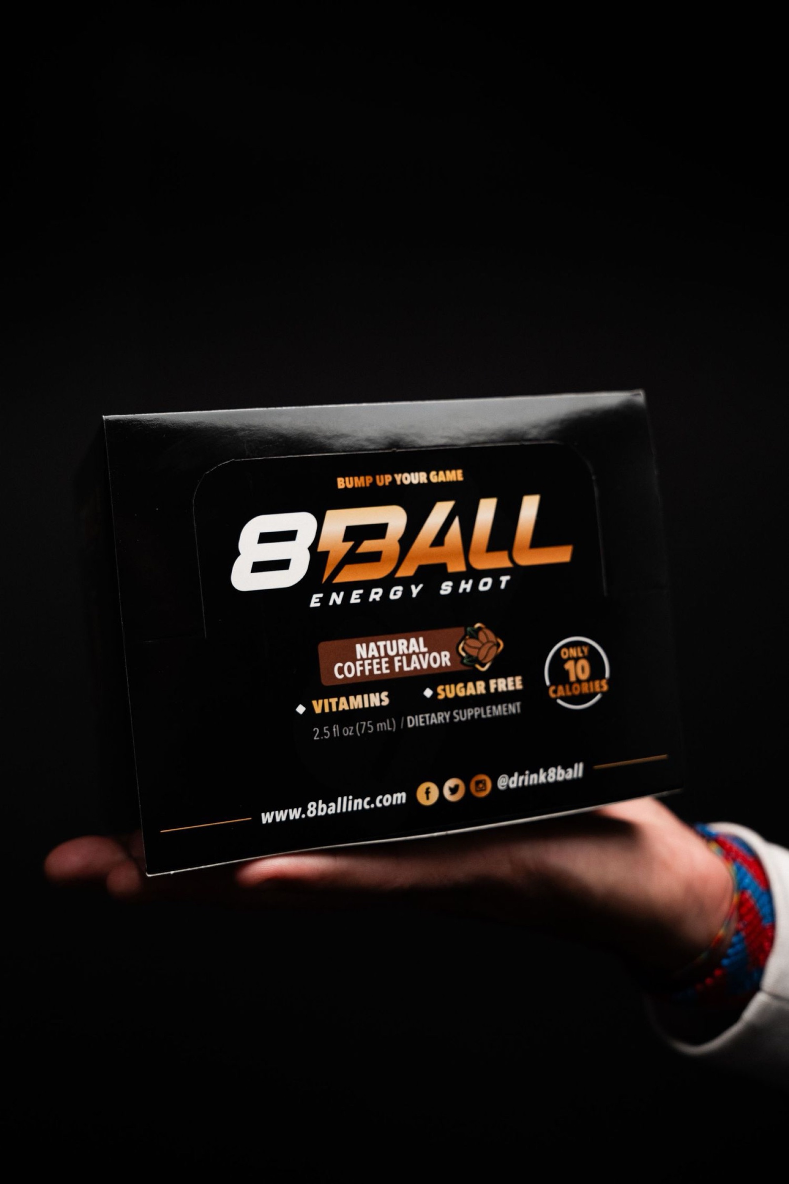 A hand holding a black 8Ball Energy Shot retail carton against a pure black background, the front panel for the coffee flavor fully lit