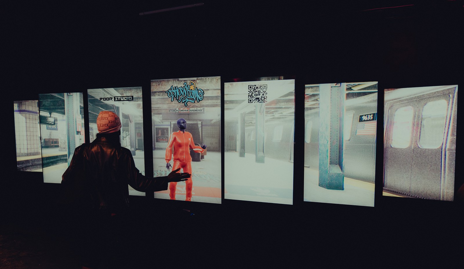 A visitor reaches toward tall screens showing a subway platform and a figure in orange in a dark room