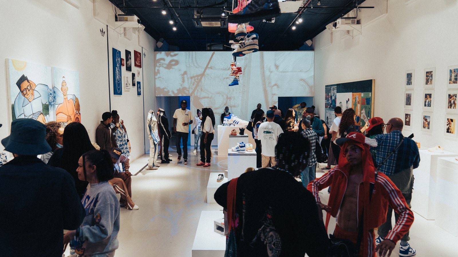 Guests mingle in a white gallery hung with paintings, sneakers on plinths, and a projected mural on the far wall