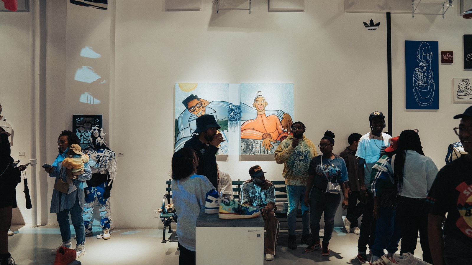 Guests mingle in a white gallery space with large cartoon murals and an Adidas trefoil logo on the wall