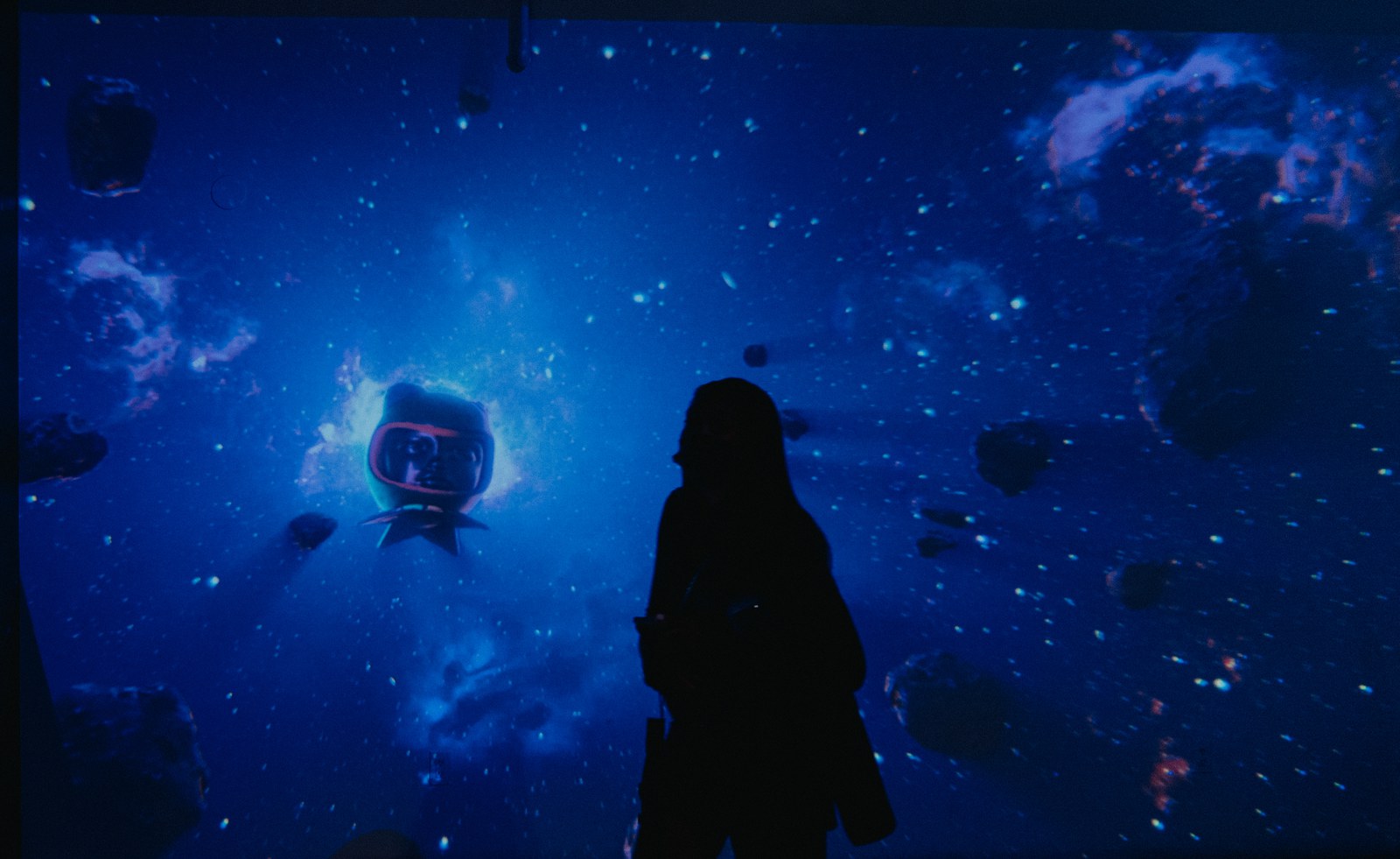Silhouette of a person facing a wall projection of blue space with an astronaut and floating asteroids
