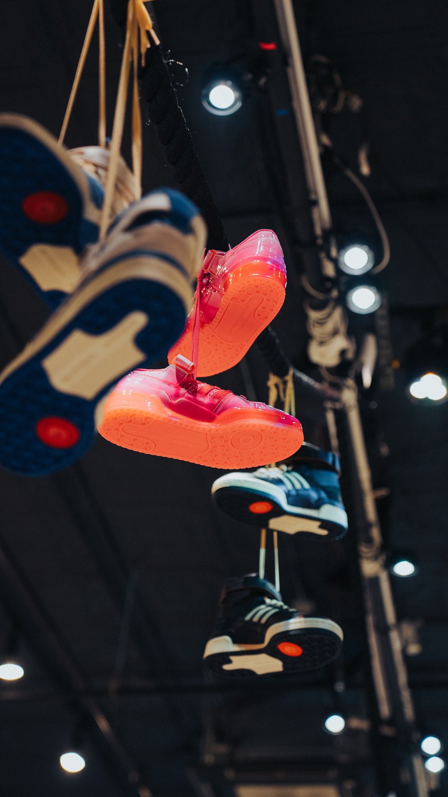 Sneakers in blue, white and glossy orange hang from cords beneath dark ceiling rigging and spotlights