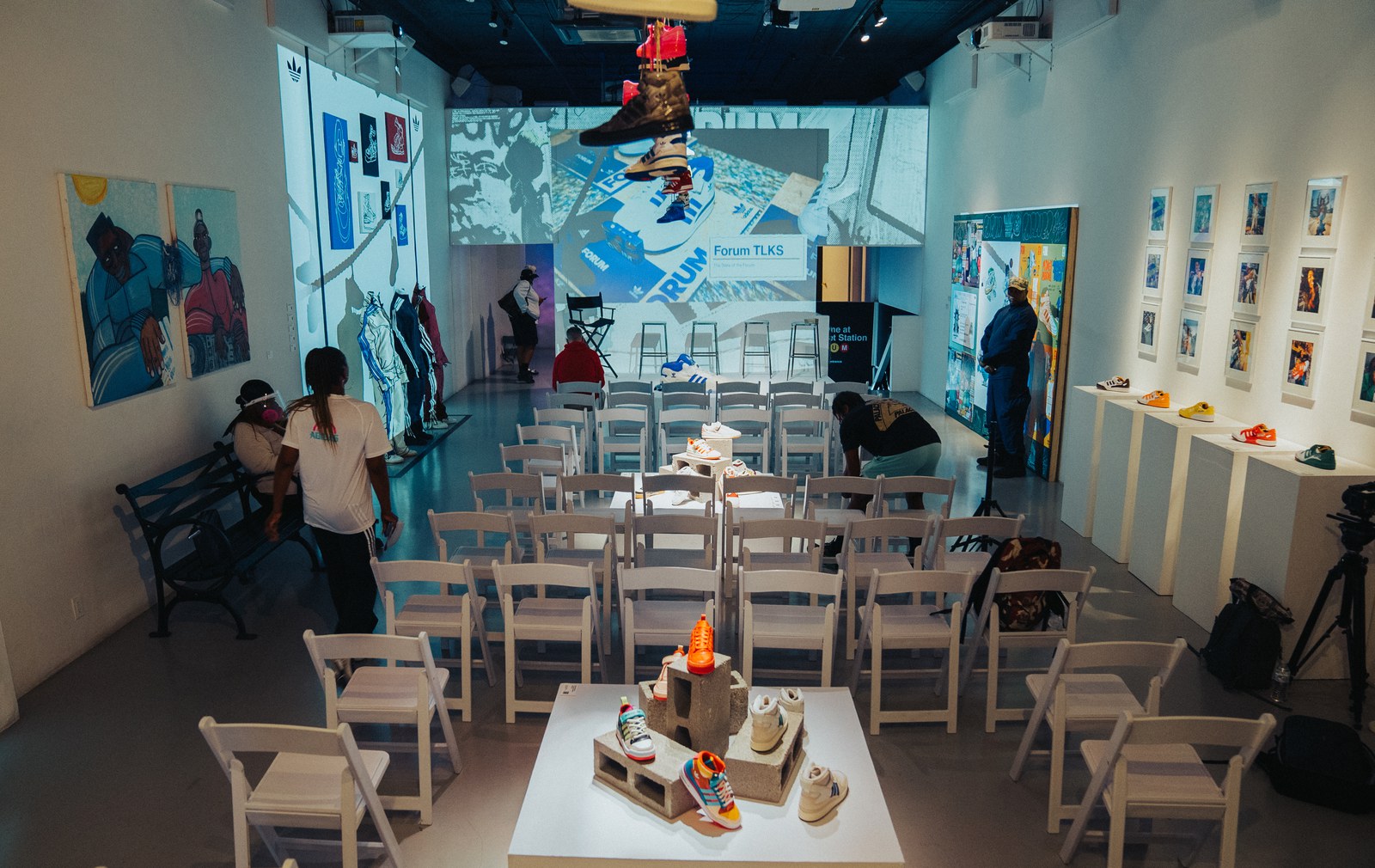 Rows of white folding chairs face a projection wall in a gallery, sneakers displayed on a low platform in front