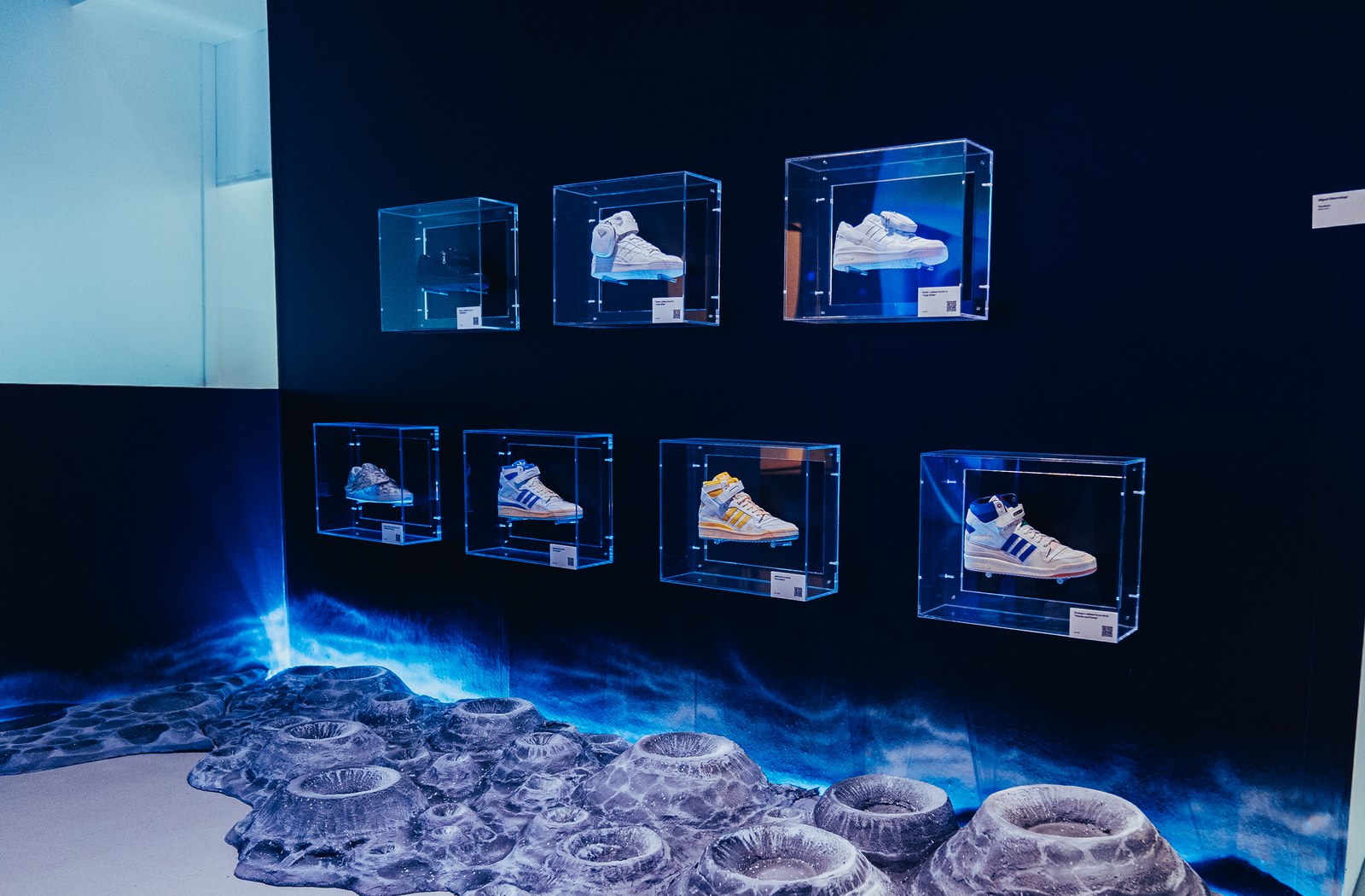 Seven clear acrylic cases holding white and blue Adidas Forum sneakers on a dark wall above a grey lunar floor