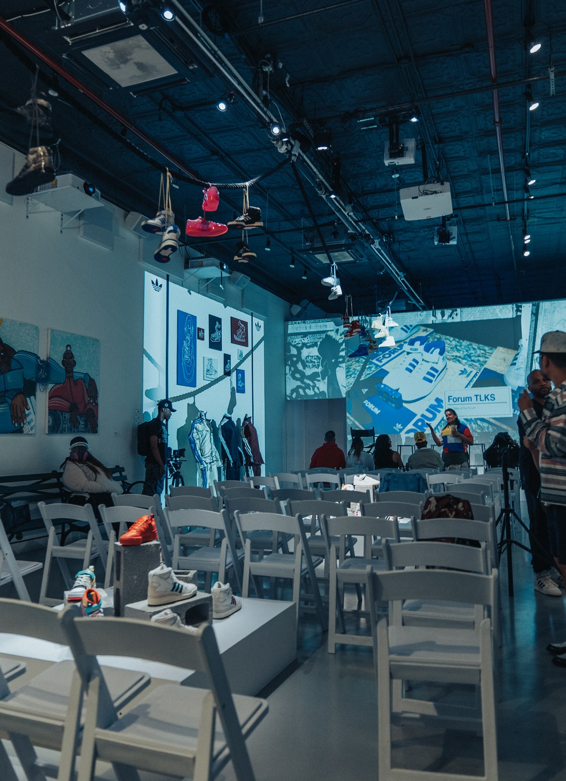 Rows of white folding chairs face a projected screen in a blue lit studio with sneakers hanging overhead