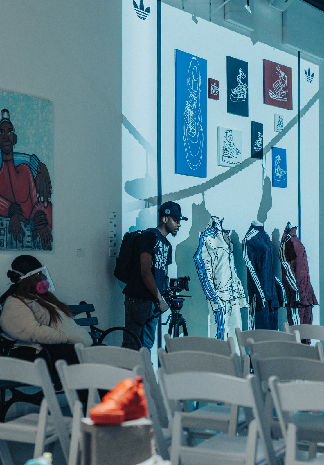 A videographer films Adidas trefoil tracksuit sketches on a white wall beside framed shoe drawings