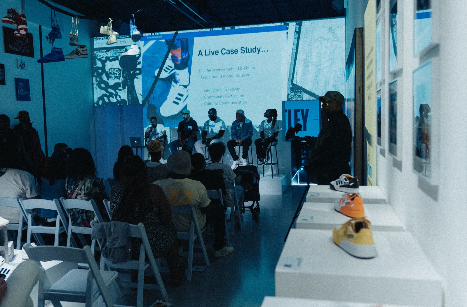 An audience in white chairs watches a panel beneath a blue projected slide, sneakers displayed on a lit shelf