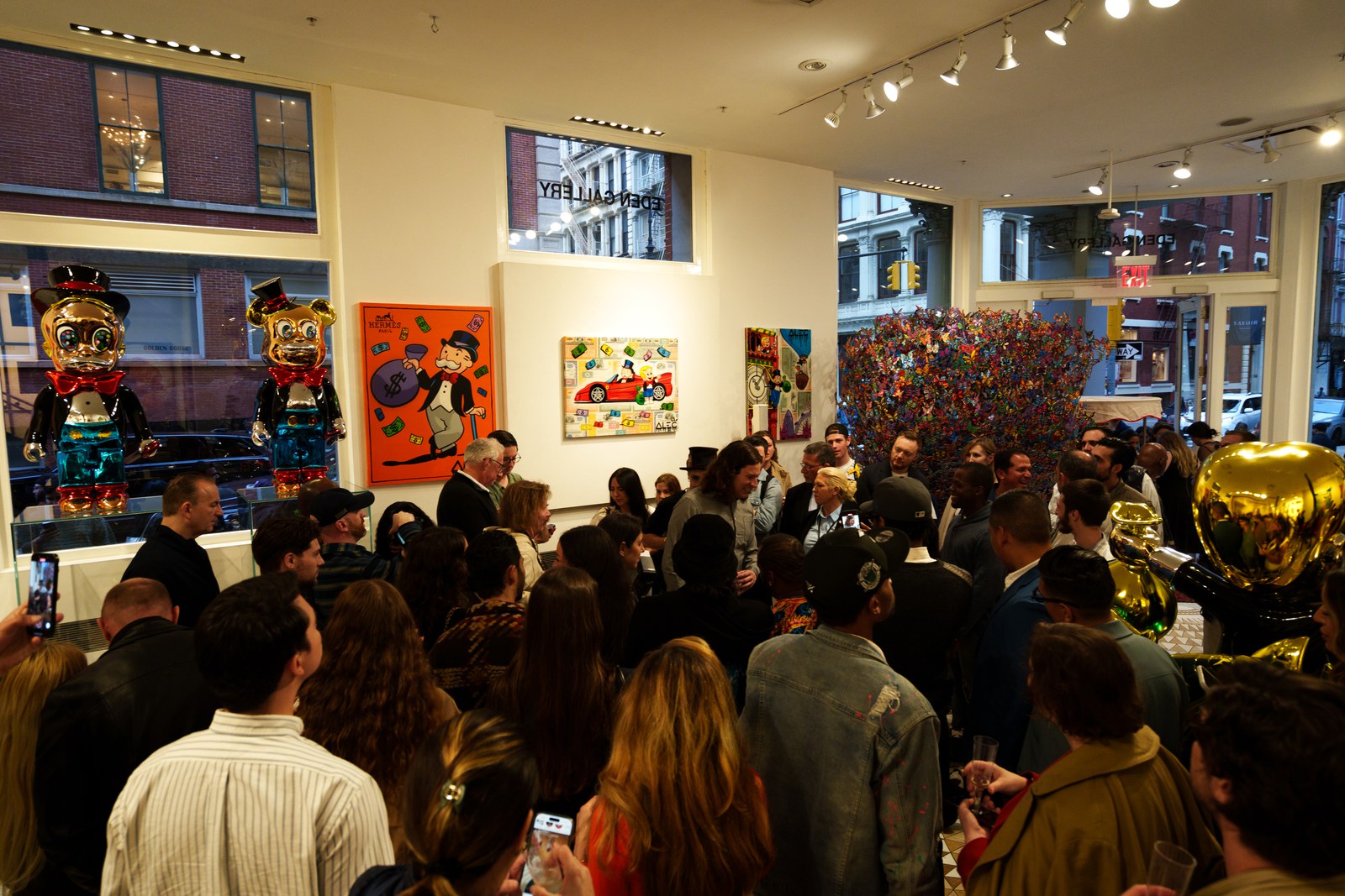 A crowd of guests fills a gallery lined with colourful cartoon paintings and Monopoly figure sculptures near tall windows