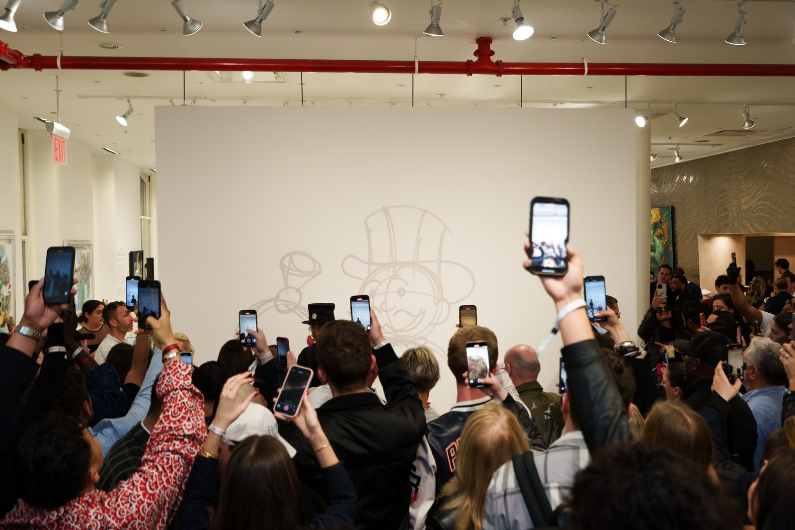 Crowd raising phones toward a line drawing of a top hatted cartoon character on a white gallery wall