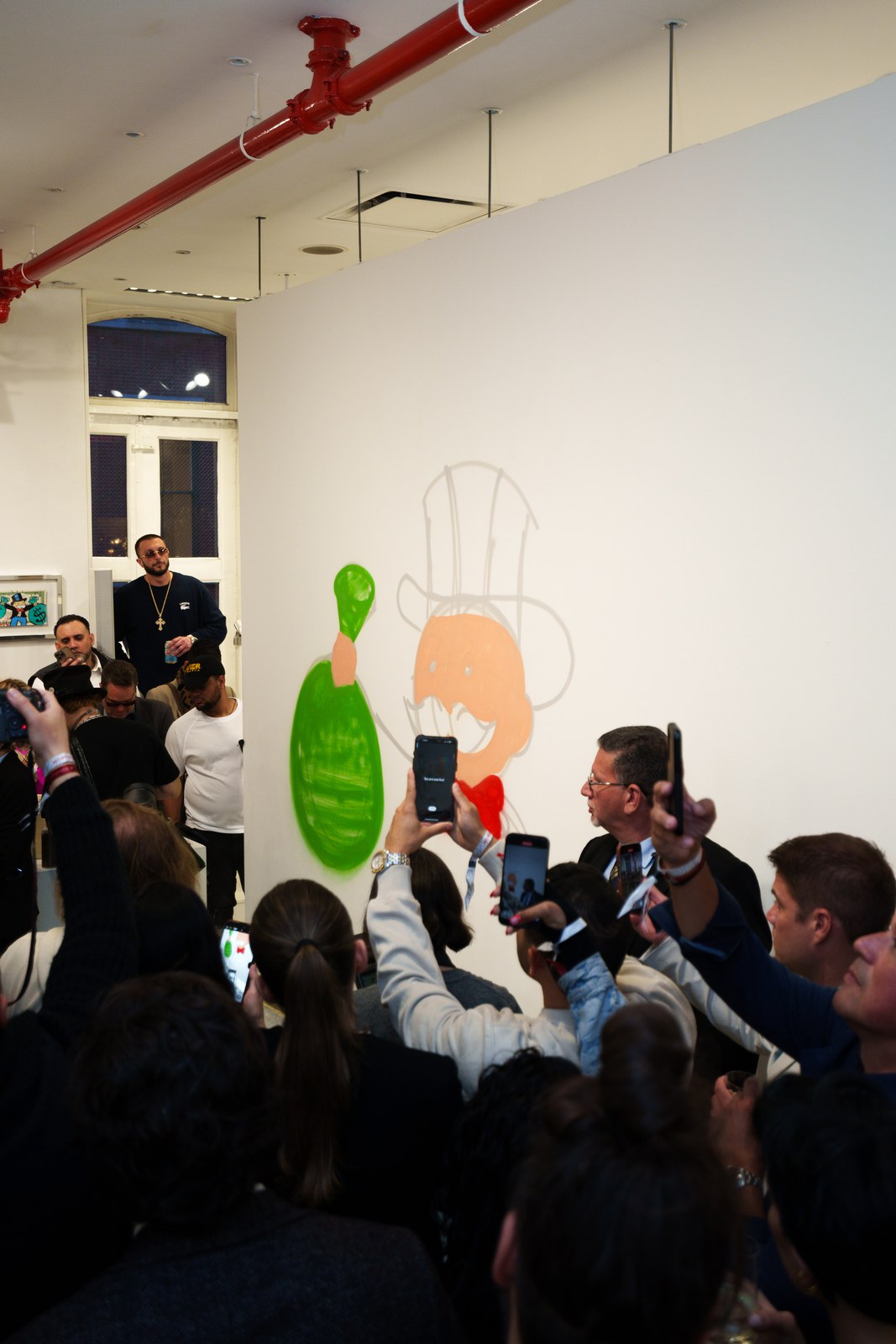 A crowd raises phones toward an artist spray painting a green and orange cartoon figure on a white gallery wall