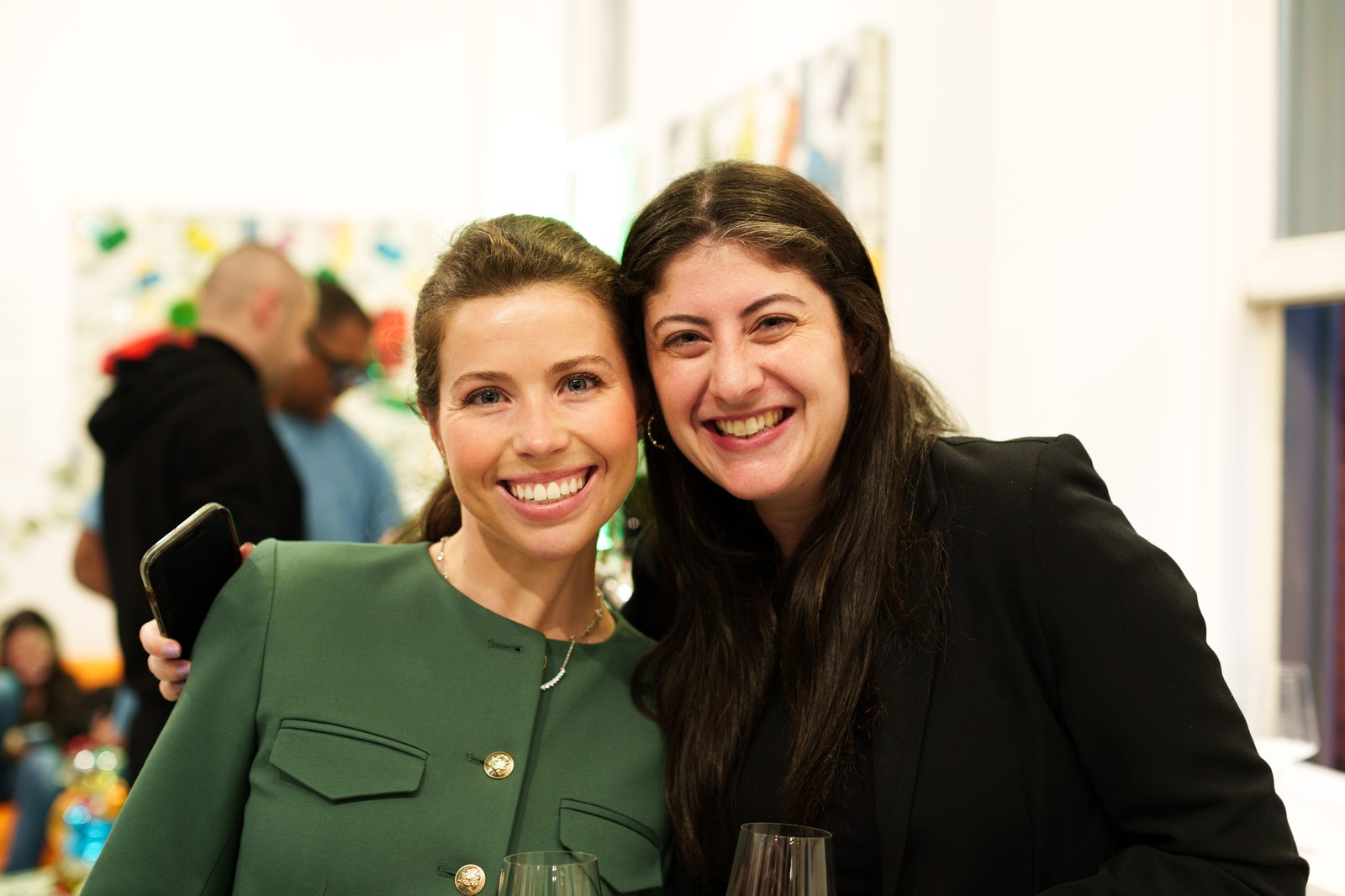 Two smiling women pose close together holding wine glasses in front of a wall of colourful artwork