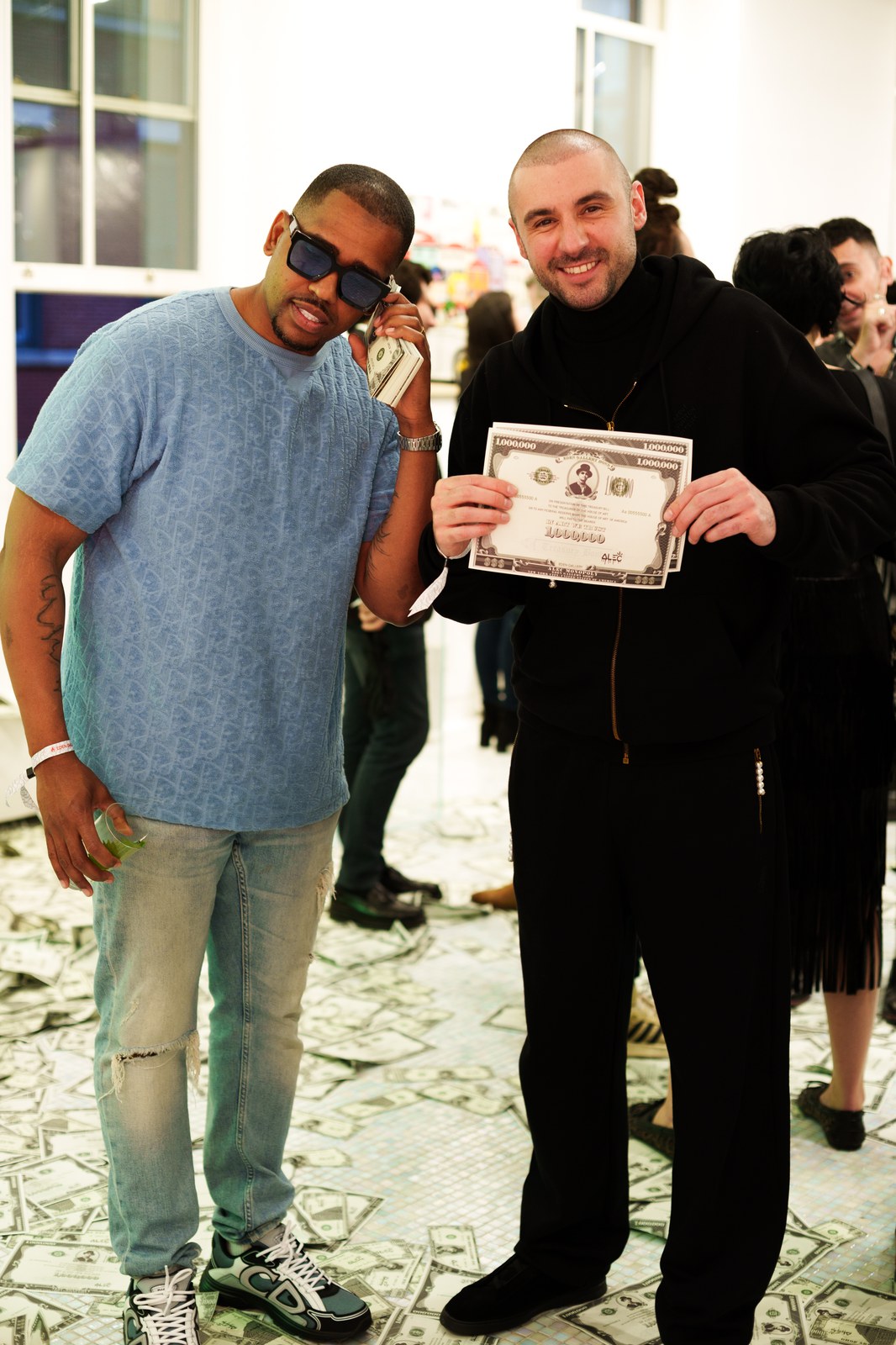 Two men posing on a floor scattered with dollar bills, one holding a printed banknote artwork
