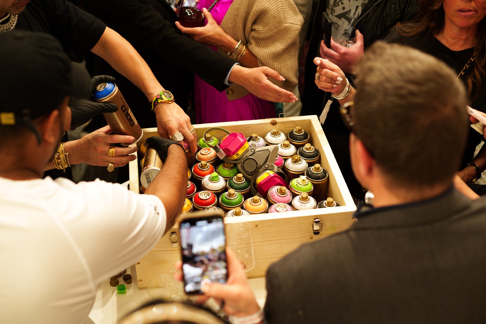 Hands reach into a wooden crate of spray paint cans while one guest films with a phone from above