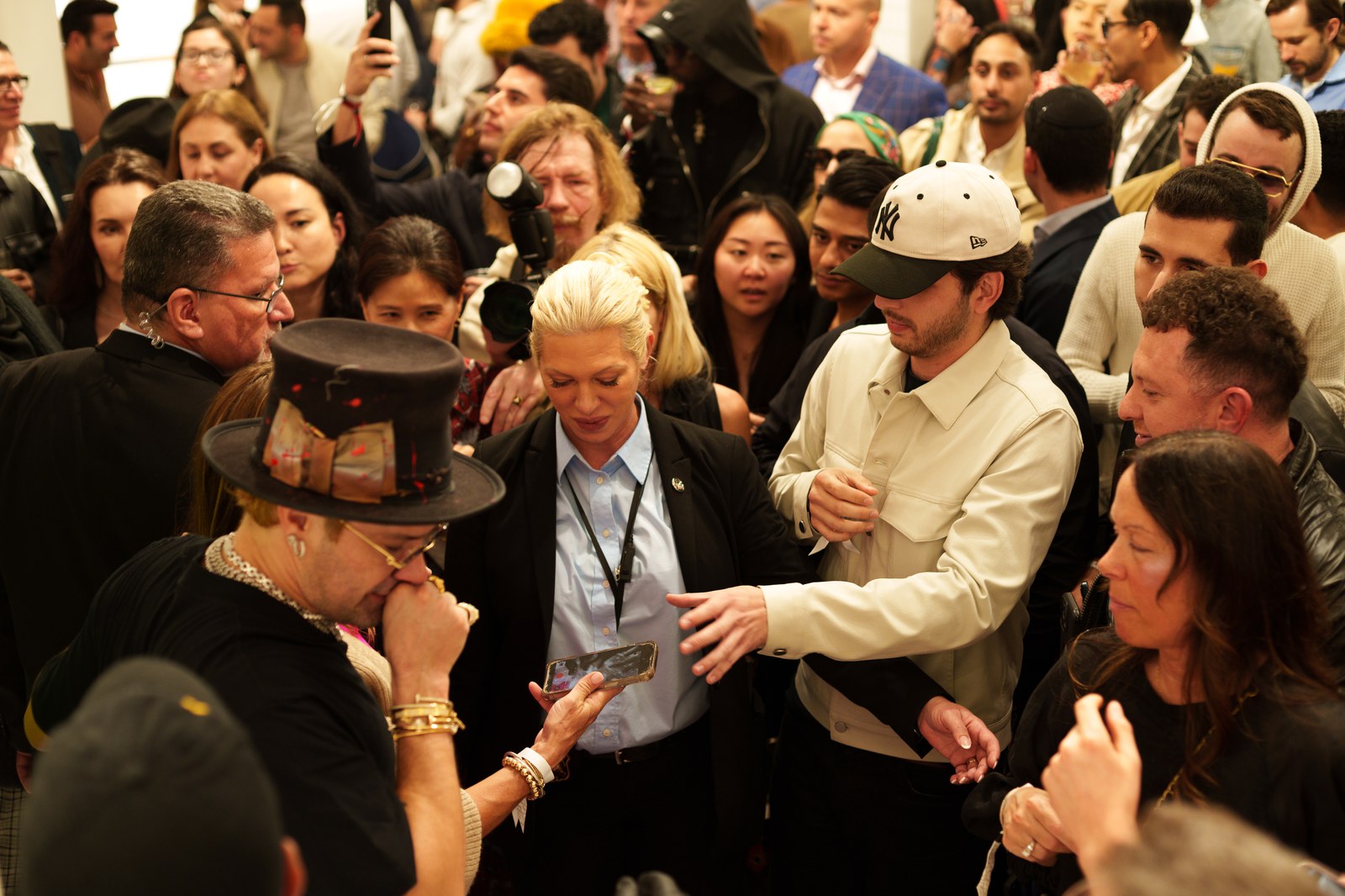 A crowd surrounds a man in a top hat as guests hold up phones and cameras in a packed gallery