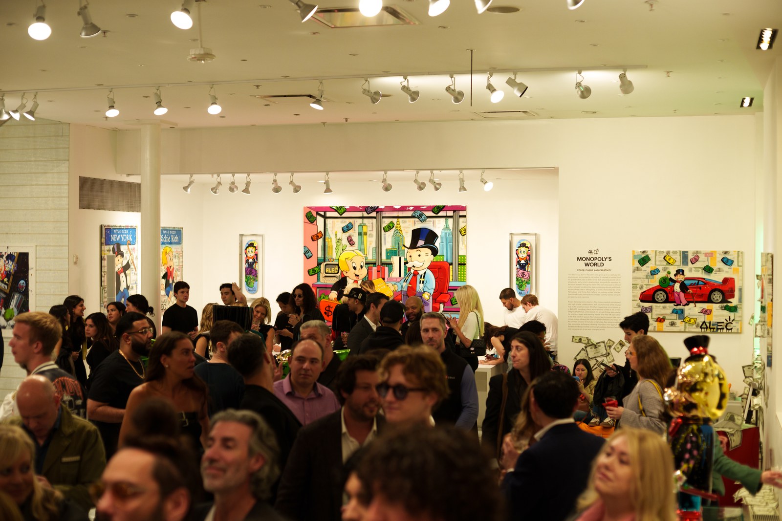 Packed gallery crowd beneath track lighting with colourful cartoon paintings on the white walls
