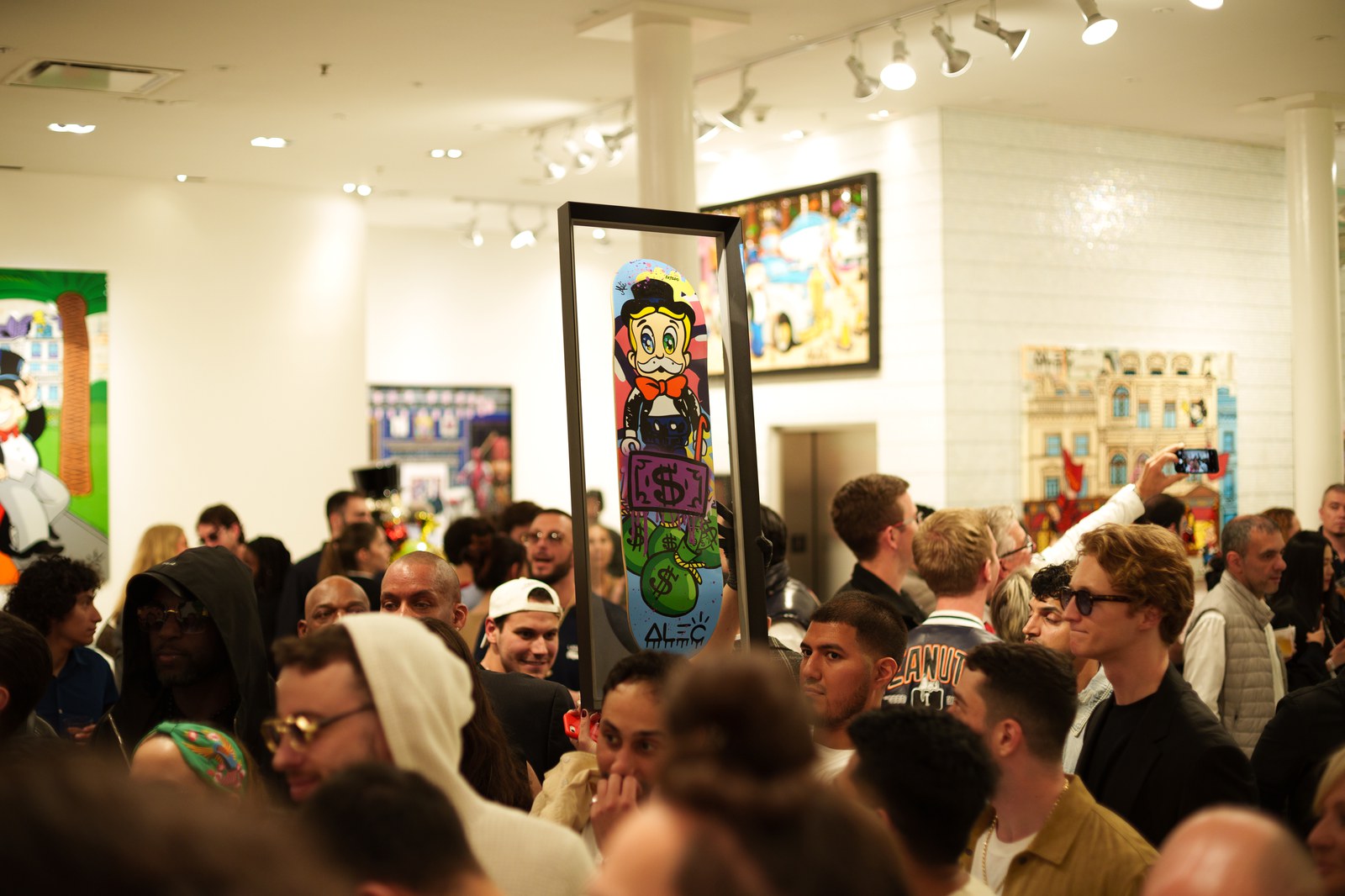 A crowded gallery gathers around a tall framed painting of a cartoon figure in a top hat
