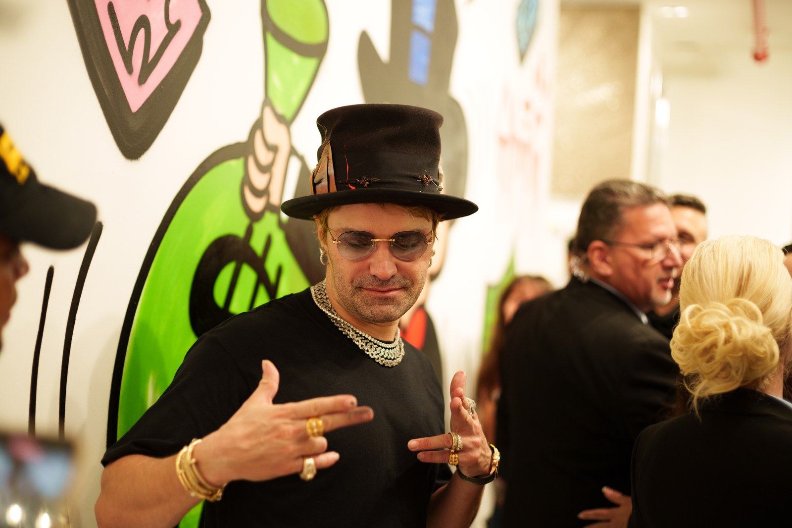 Artist in black top hat and tinted glasses poses by colorful graffiti at Eden Gallery