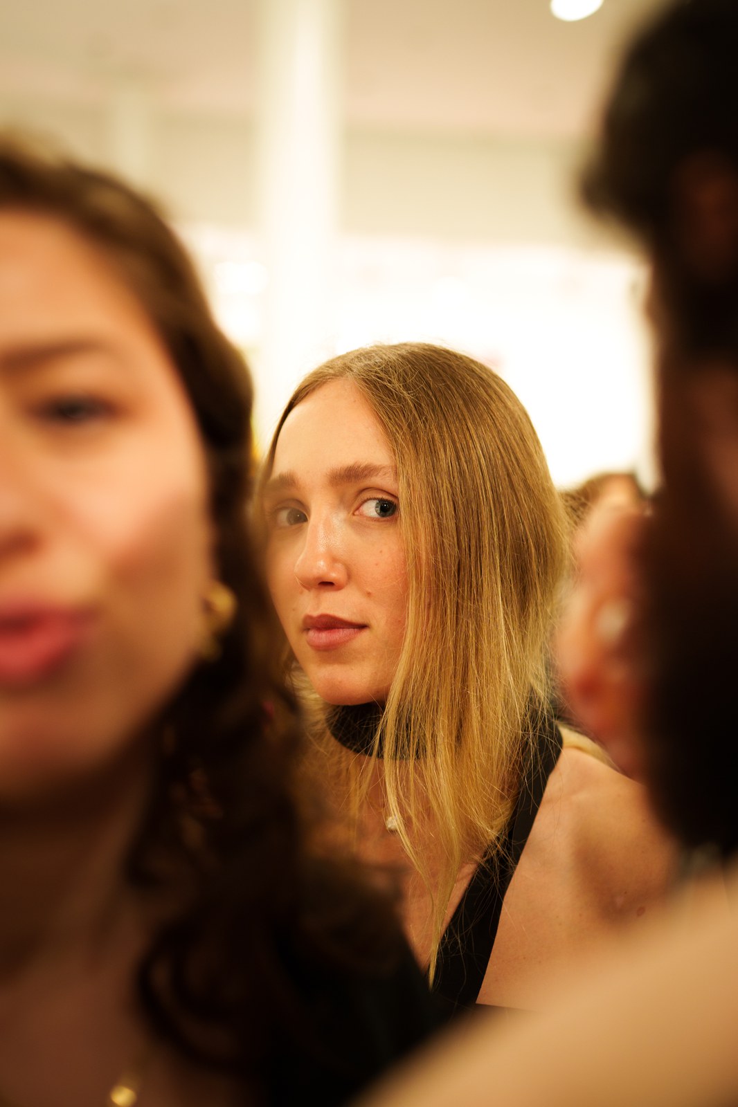 Young woman with long blonde hair glancing sideways between two blurred guests in a bright gallery