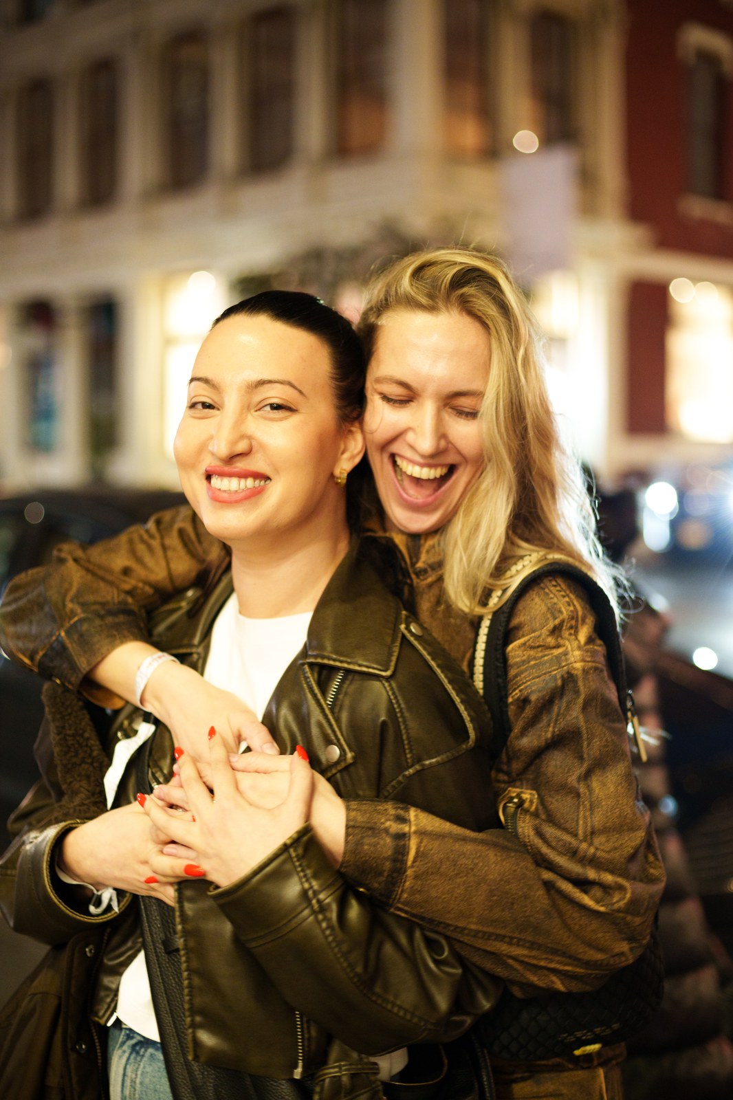 Two laughing women in leather jackets hug on a city street at night, blurred storefront lights behind them