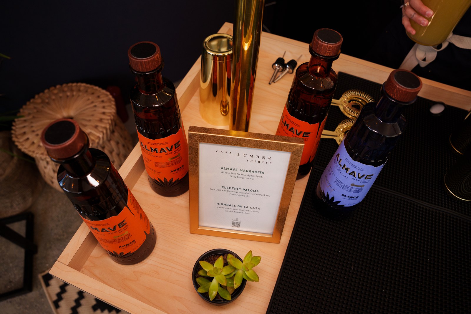 Almave Blanco and Ambar bottles surround a framed Casa Lumbre cocktail menu on a wooden bar cart