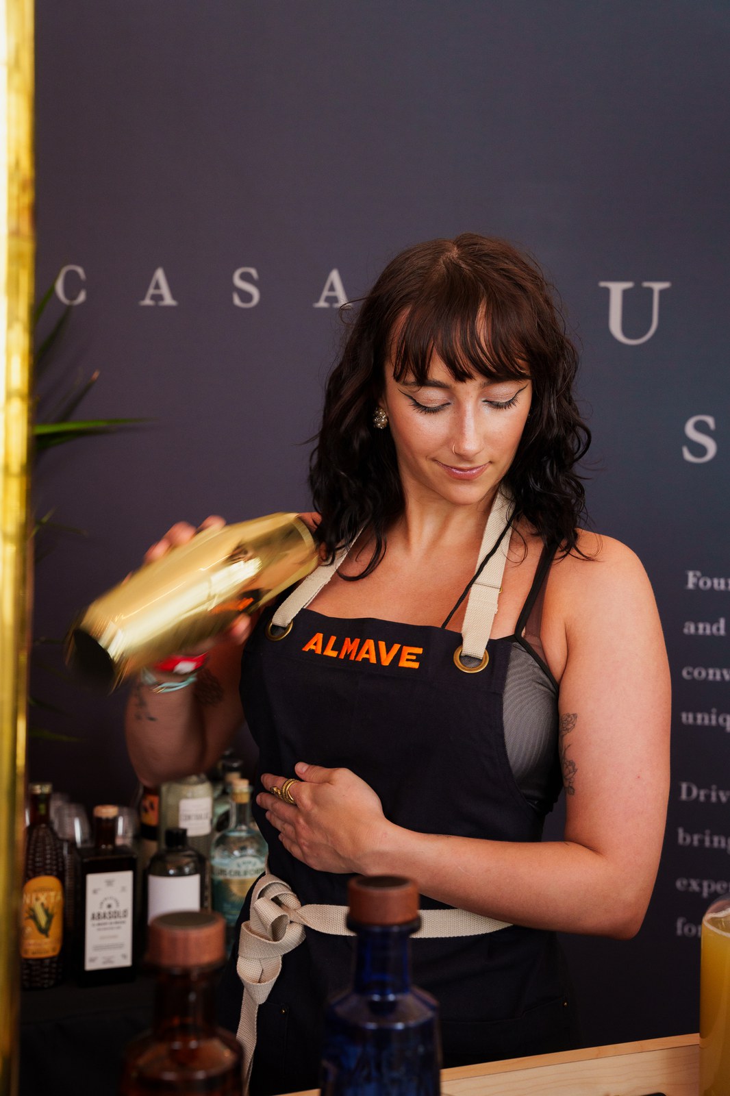 Bartender in an Almave apron shakes a gold cocktail shaker at the Casa Lumbre bar