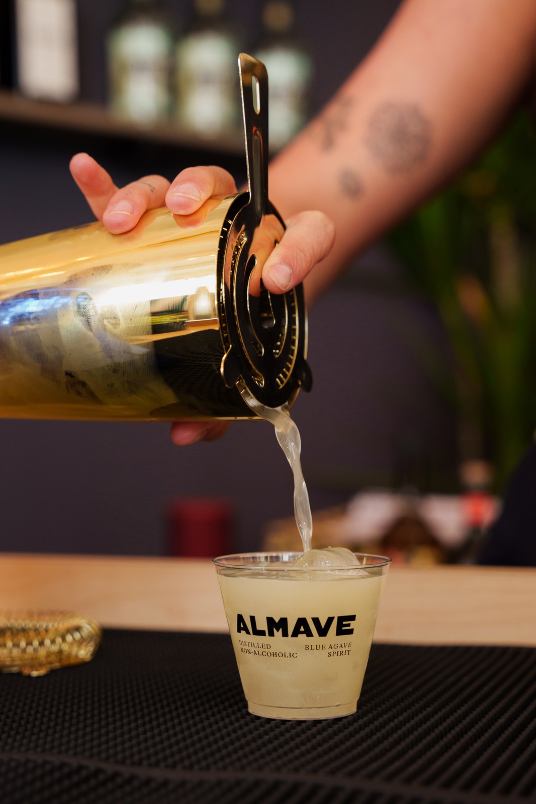 Strained agave drink is poured from a gold shaker into a branded Almave cup with ice