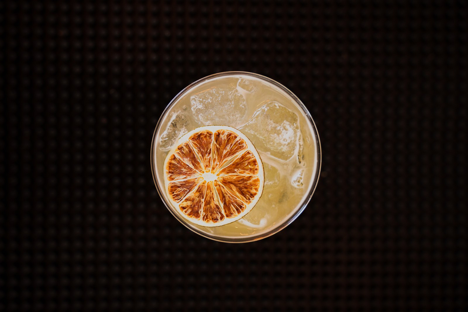 Overhead view of an iced agave cocktail topped with a dried citrus wheel on a dark mat