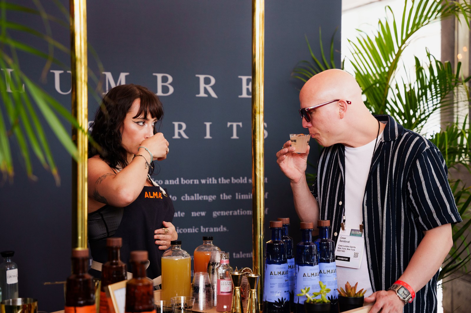 Two guests sip tasting samples at a bar counter with Almave bottles beneath a Casa Lumbre Spirits wall