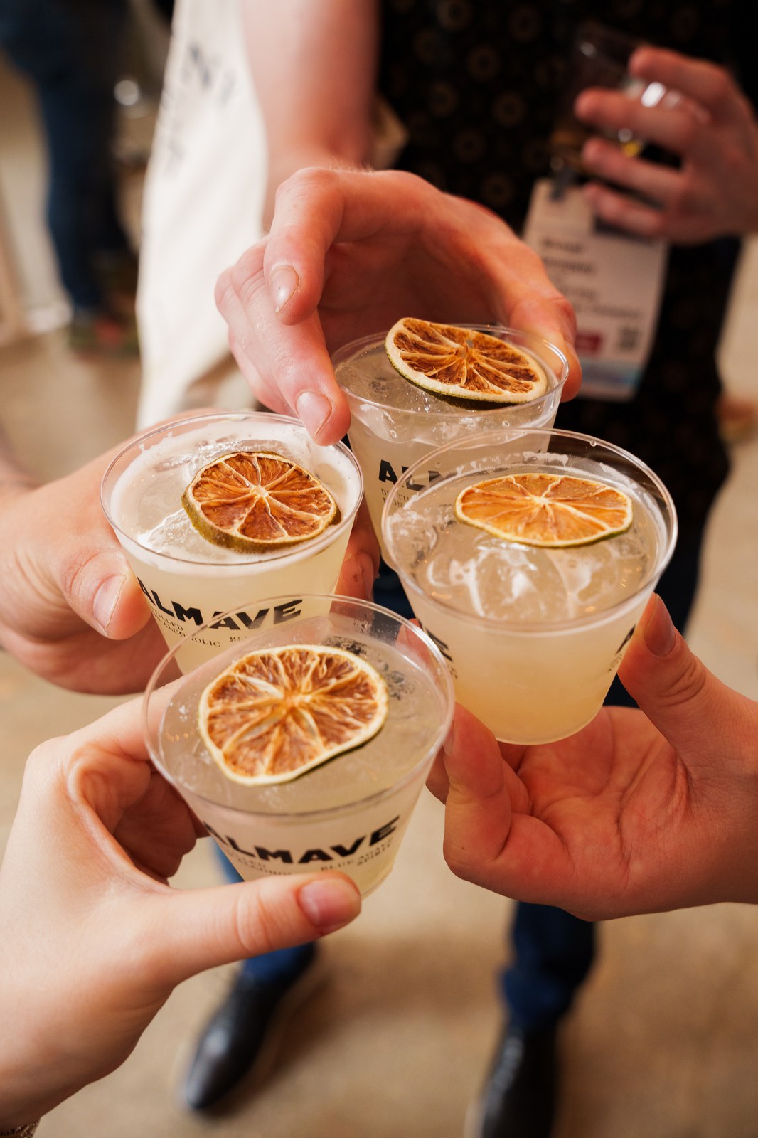 Four hands raise plastic cups of pale cocktails topped with dried lime slices, each cup printed with the Almave logo
