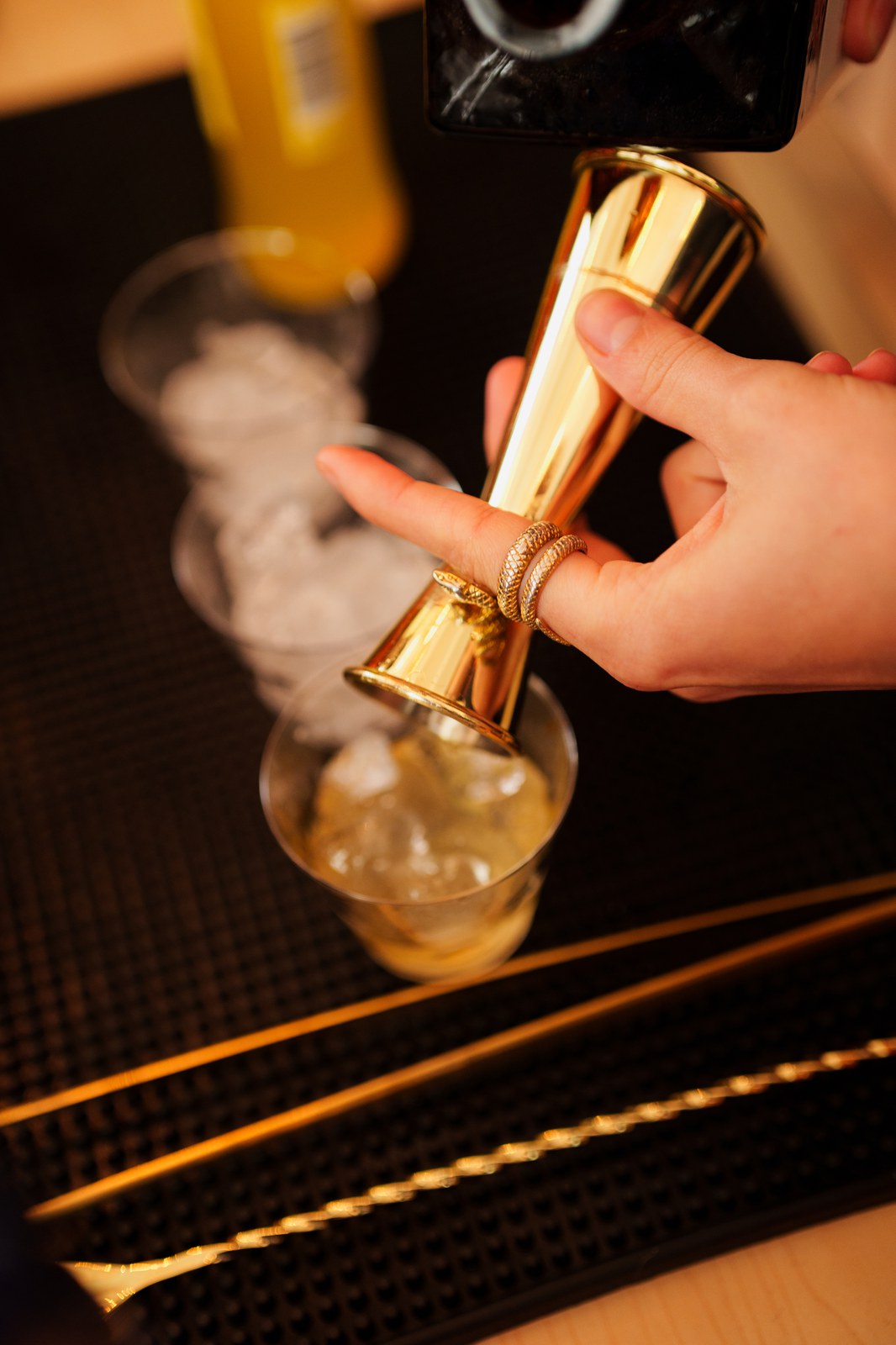 A hand pours from a gold jigger into a glass of ice on a bar mat with gold bar spoons