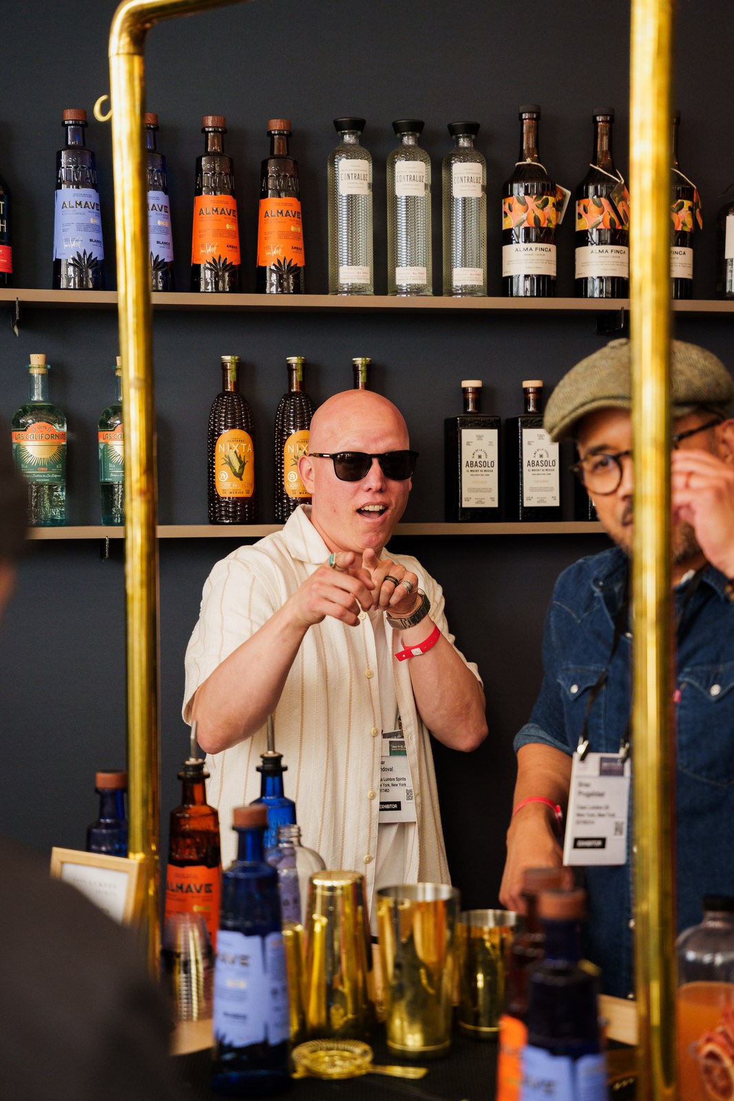 A bald bartender in sunglasses and a striped shirt points across a bar backed by shelves of Almave bottles