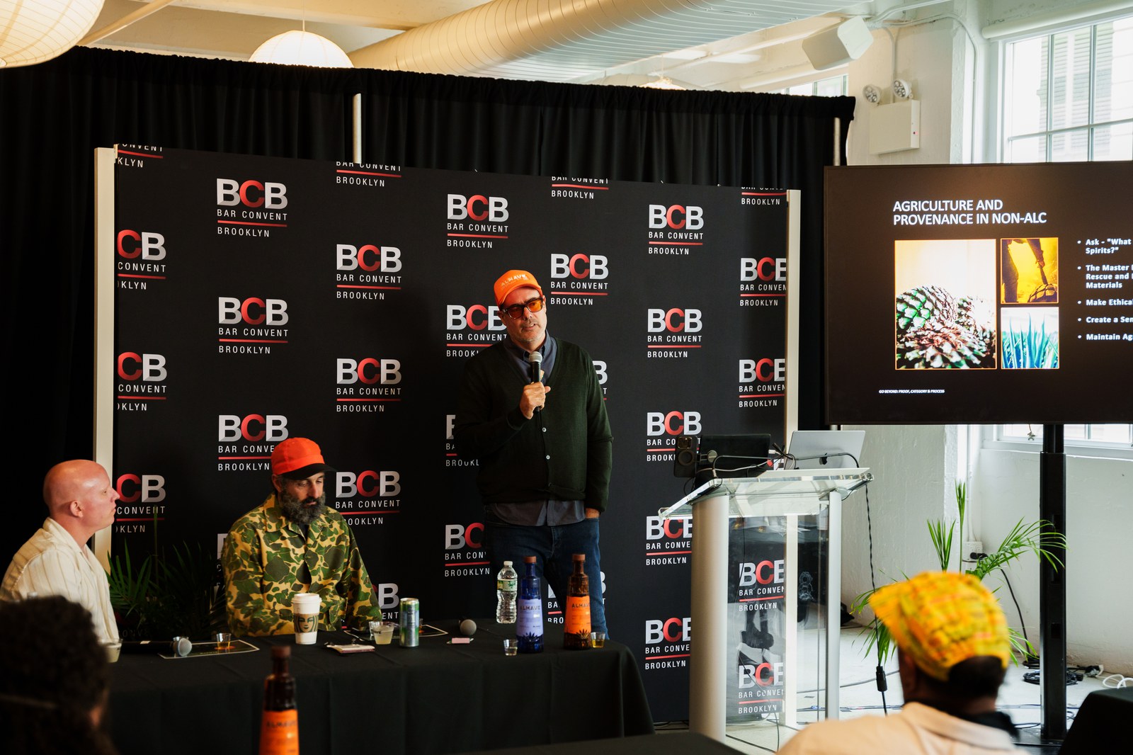 Speaker in an orange beanie addressing a panel in front of a Bar Convent Brooklyn backdrop