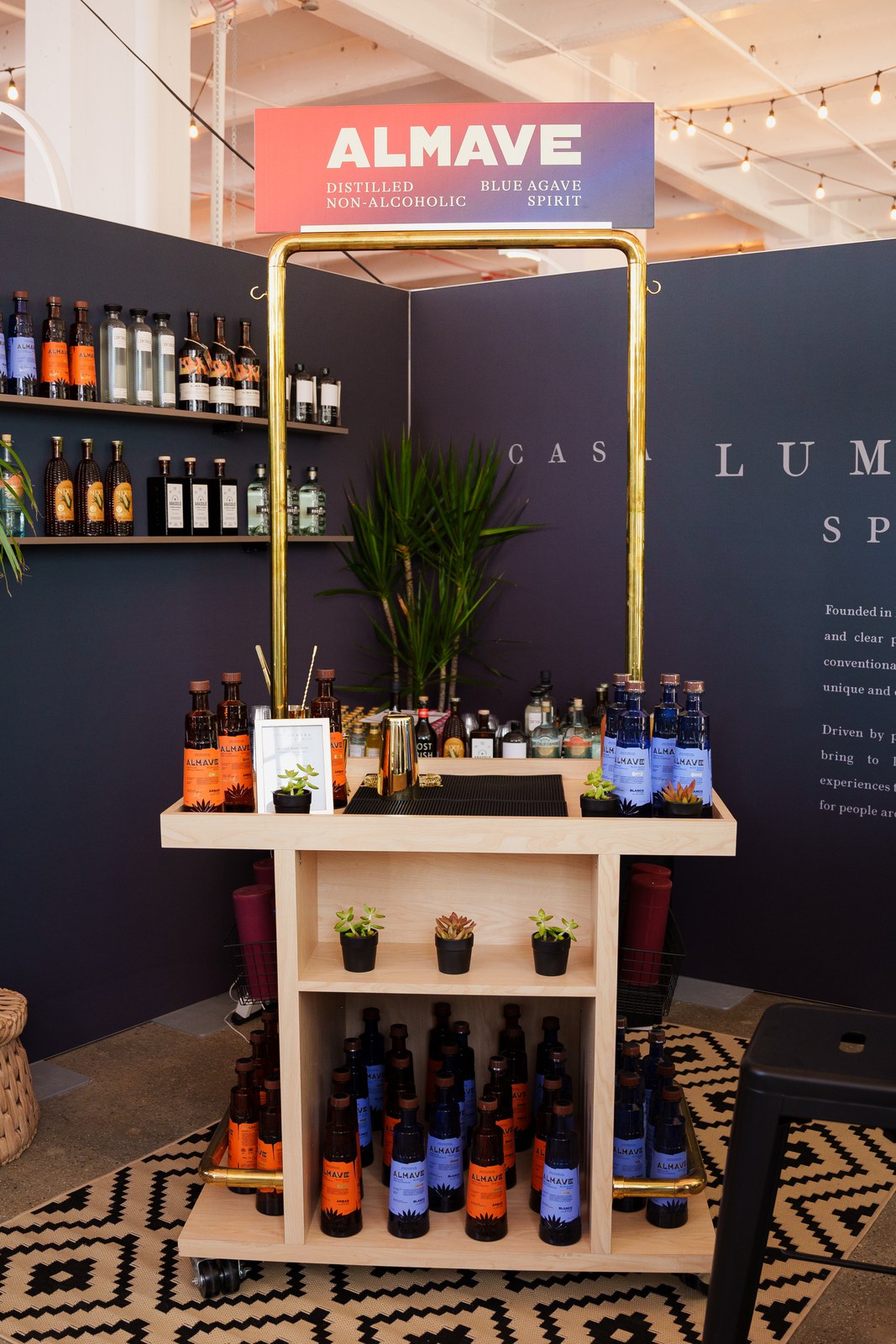 Almave branded wooden bar cart with a gold frame, potted plants and orange and blue bottles against a navy wall