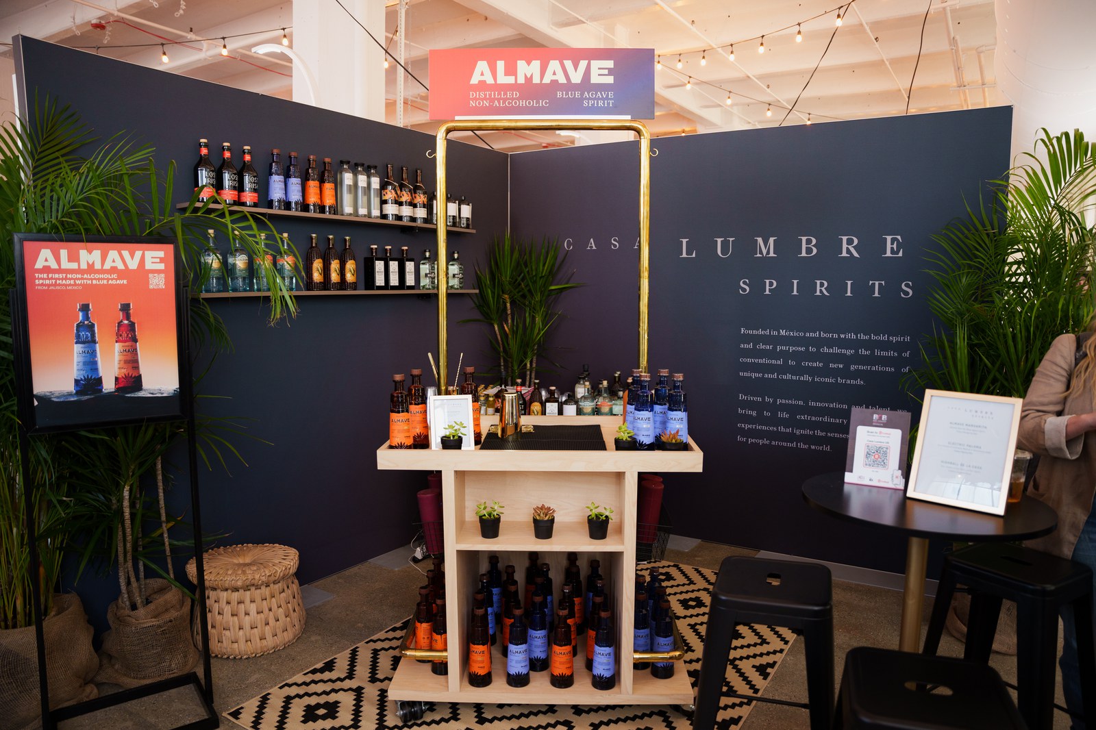 A Casa Lumbre Spirits booth with a red Almave sign, wooden shelves of bottles and potted palms