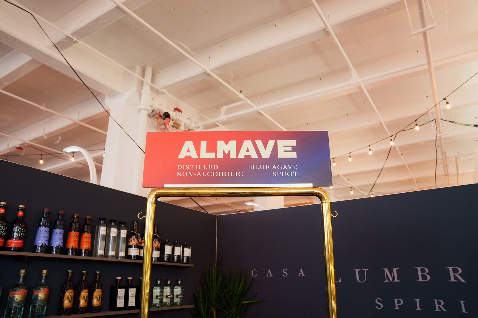 Almave sign reading distilled non alcoholic blue agave spirit above a gold framed bar shelf of bottles