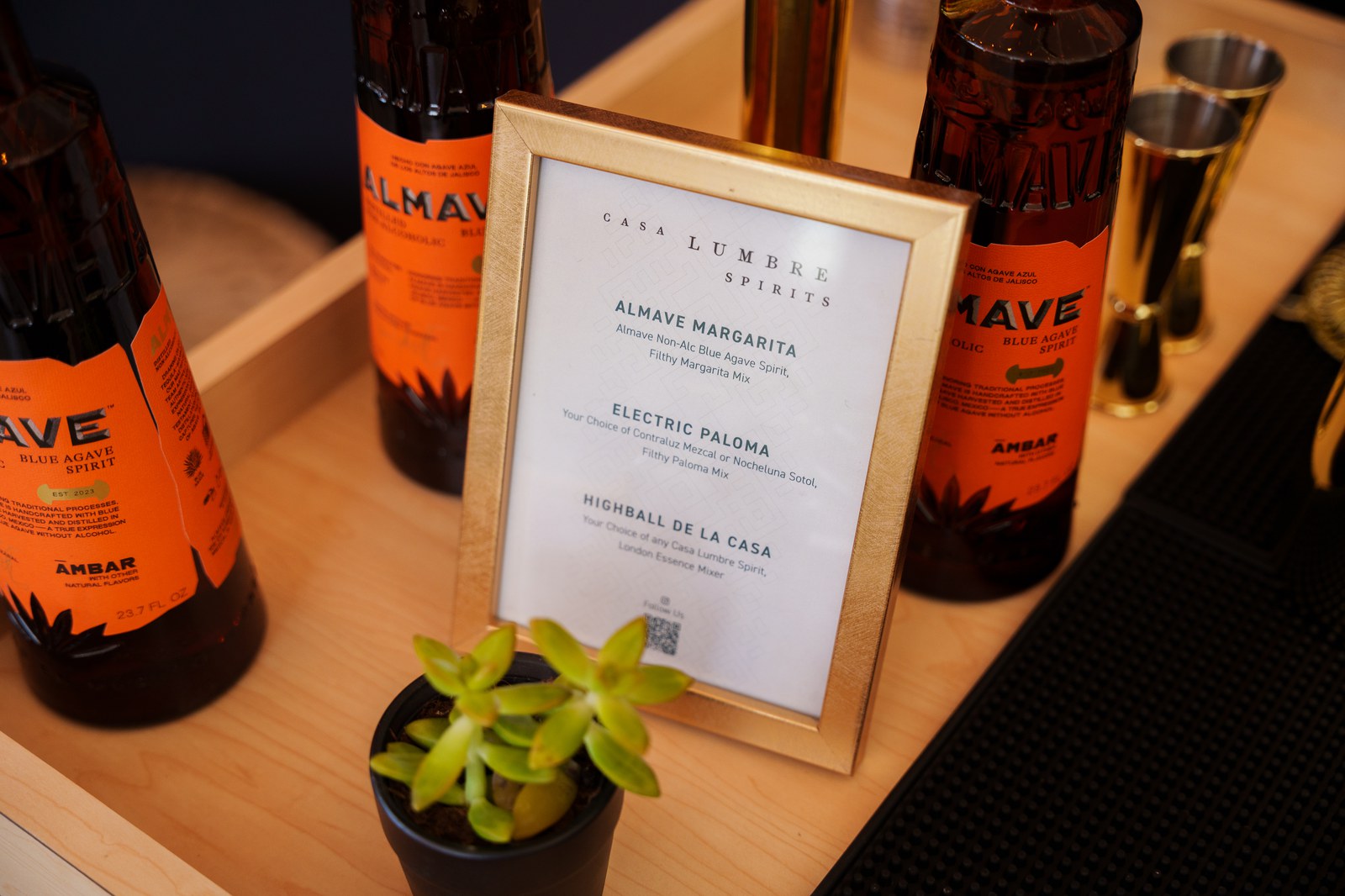 A framed Casa Lumbre cocktail menu sits in a wooden tray beside Almave bottles and a small potted succulent