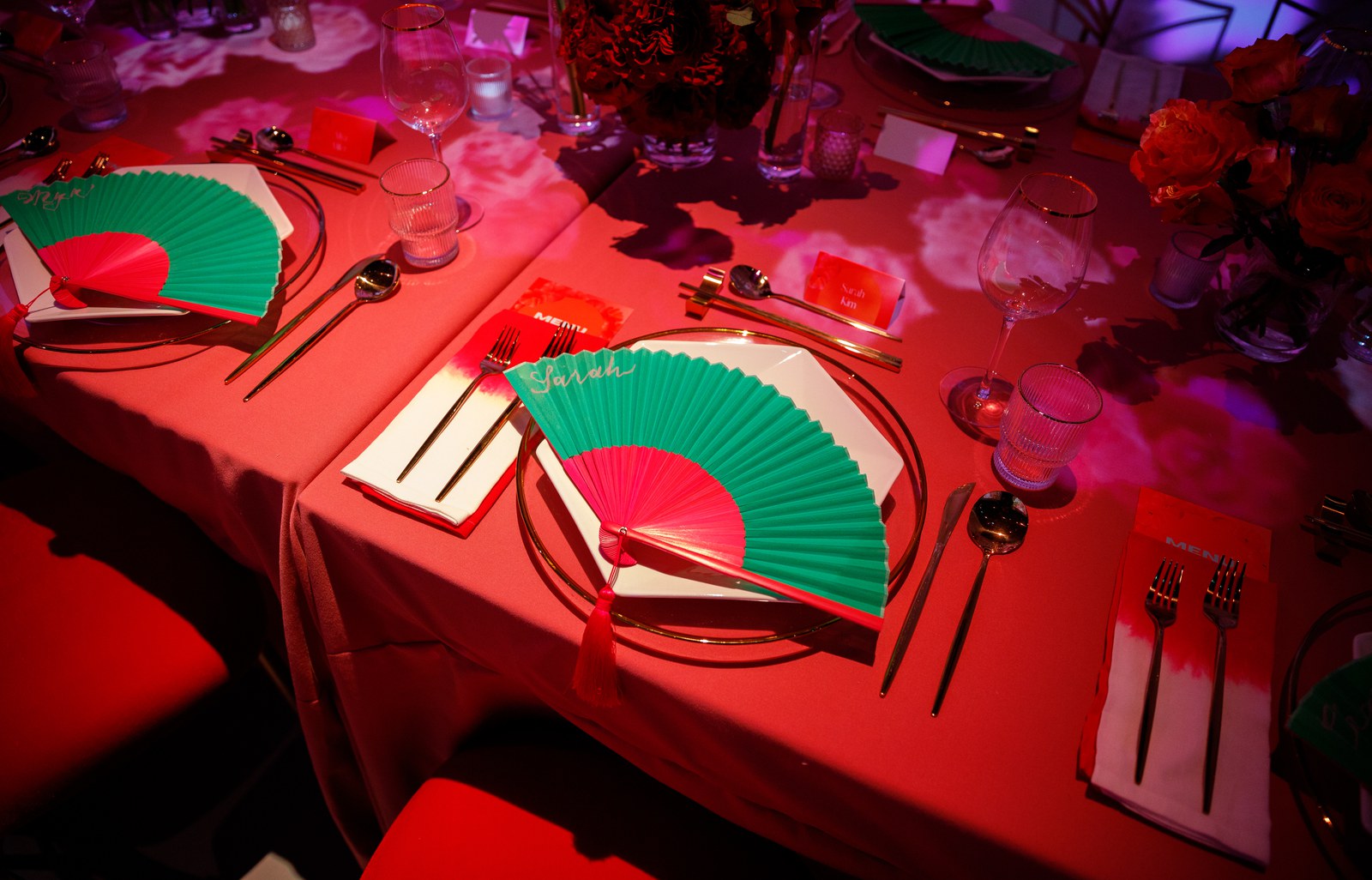 Place settings on a red tablecloth with green and pink folding fans under pink projected light