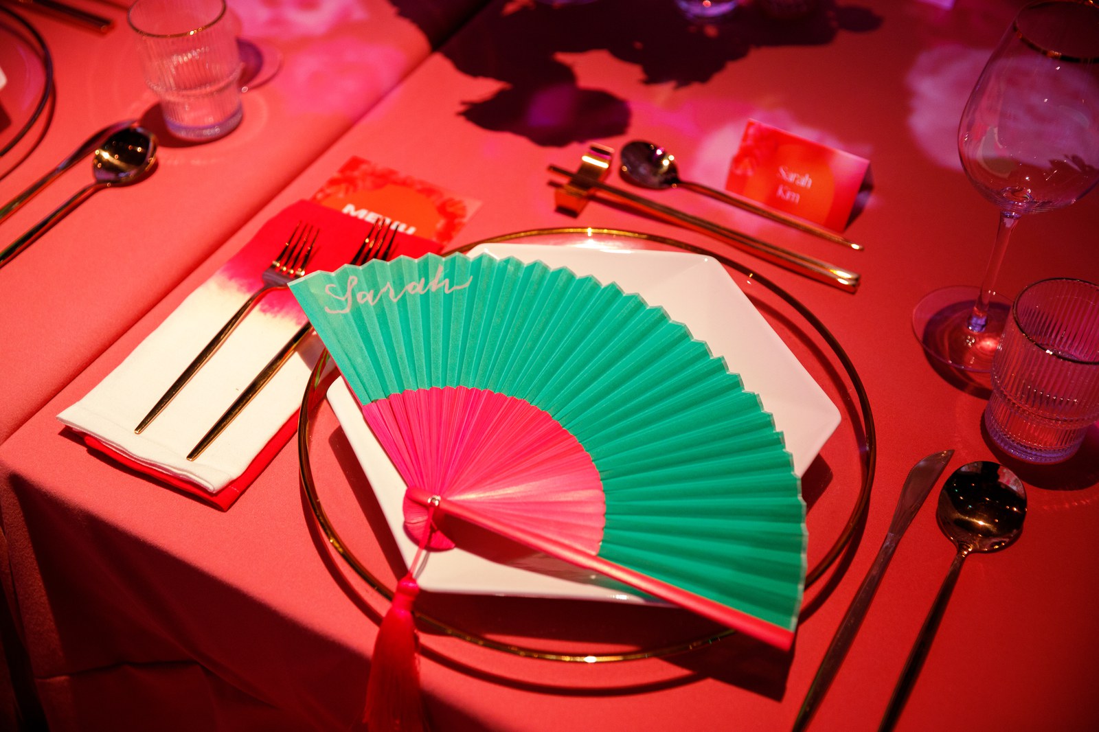 A green and pink folding fan with a handwritten name card rests on a place setting under red light