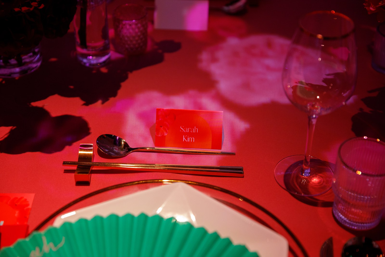 Red lit place setting with a name card, gold chopsticks and spoon, a wine glass and a green folding fan