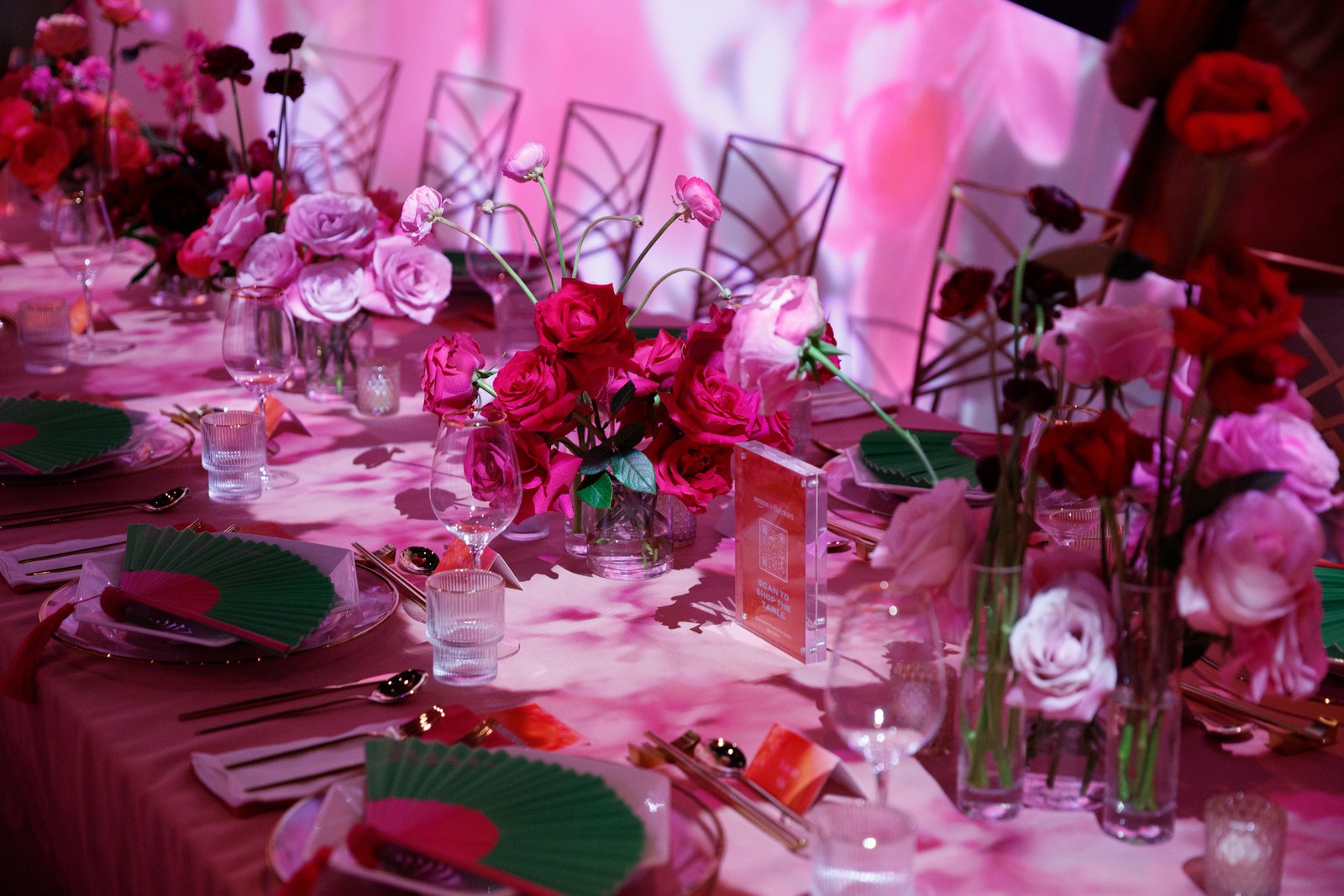 A long table set with pink and red roses, green folding fans and gold cutlery under pink projected light