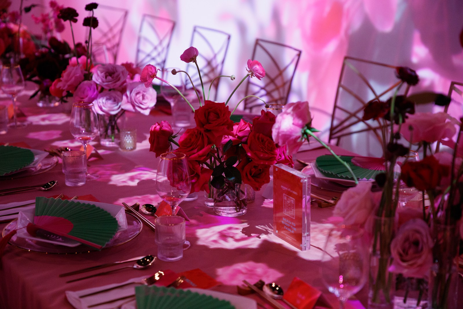 A long dinner table lined with red and pink roses, gold cutlery and folding fans under pink projected light