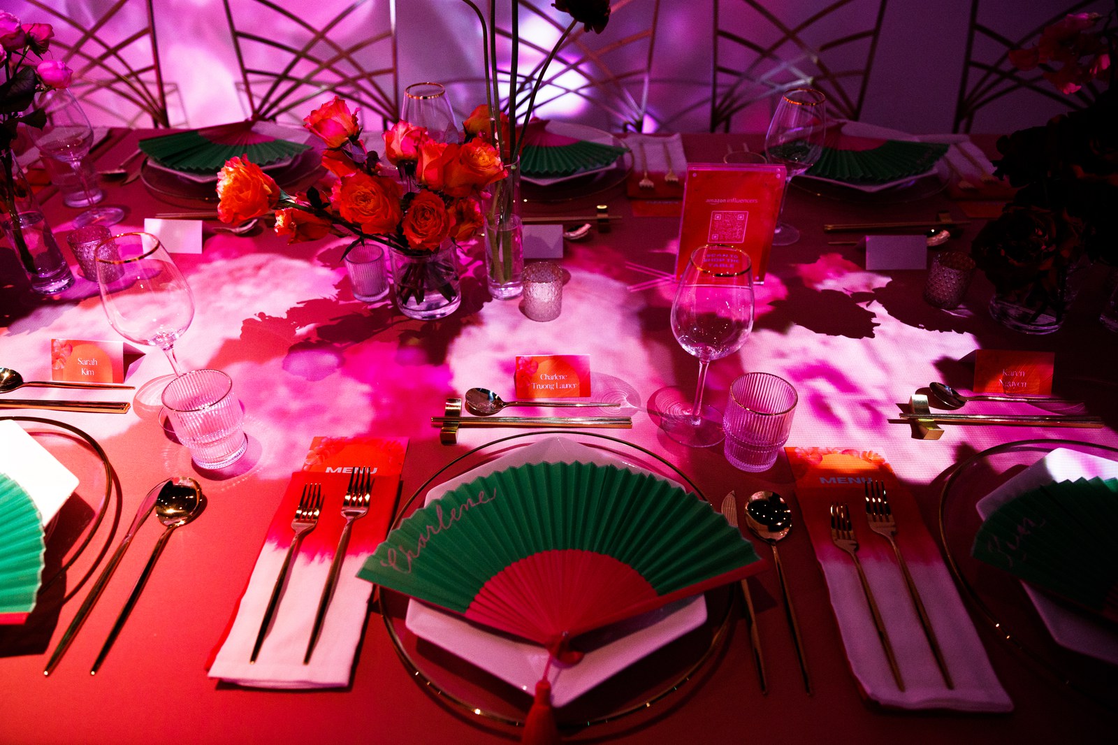 Place settings with green and red folding fans and orange menus on a red tablecloth under magenta light