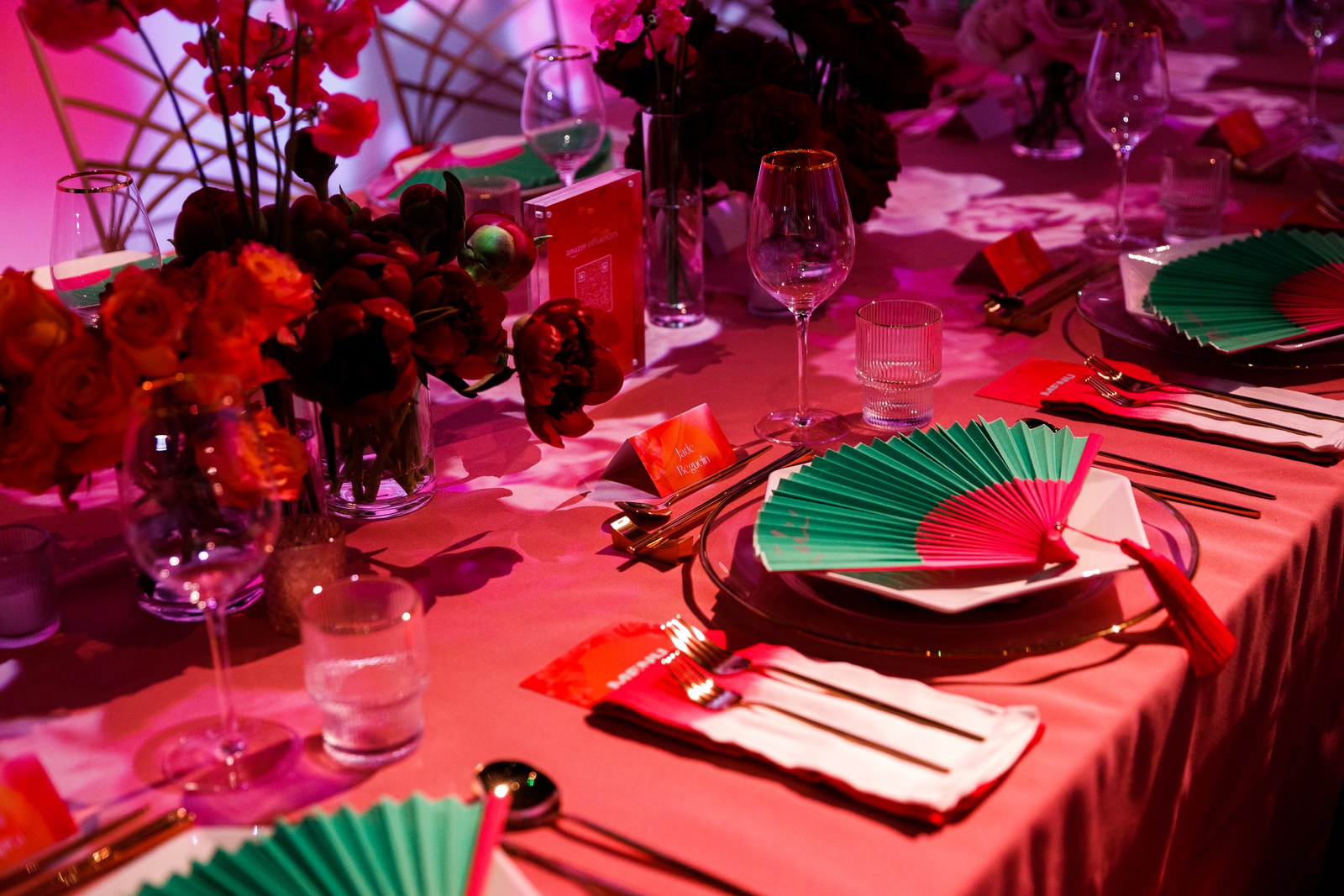 Place settings with green and red fans, red napkins and wine glasses beside bouquets of red flowers in pink light