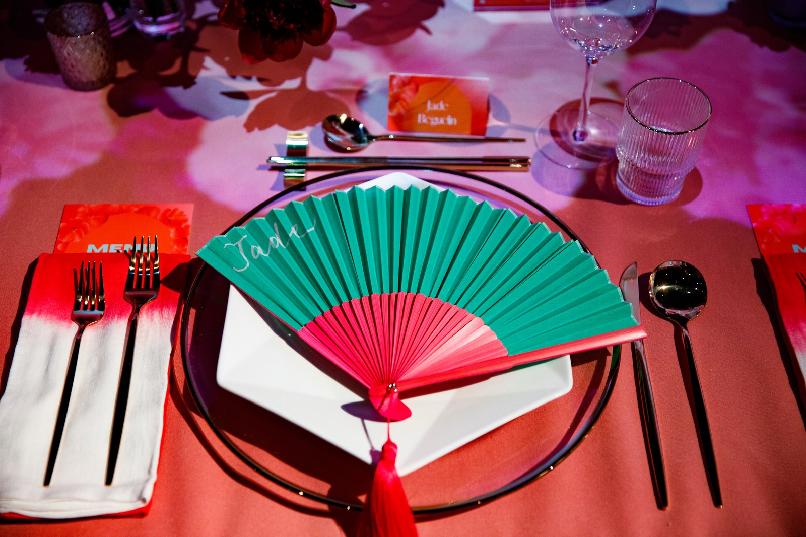 Green and red folding fan laid on a white plate on a red lit table with gold cutlery